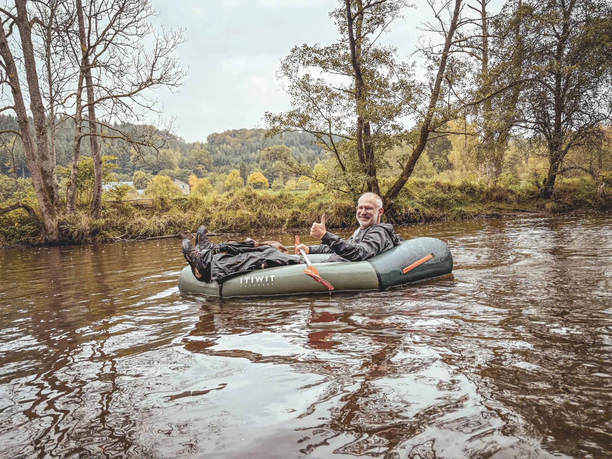 A smiling adventurer in a packraft, surrounded by lush green countryside in the Ardennes.