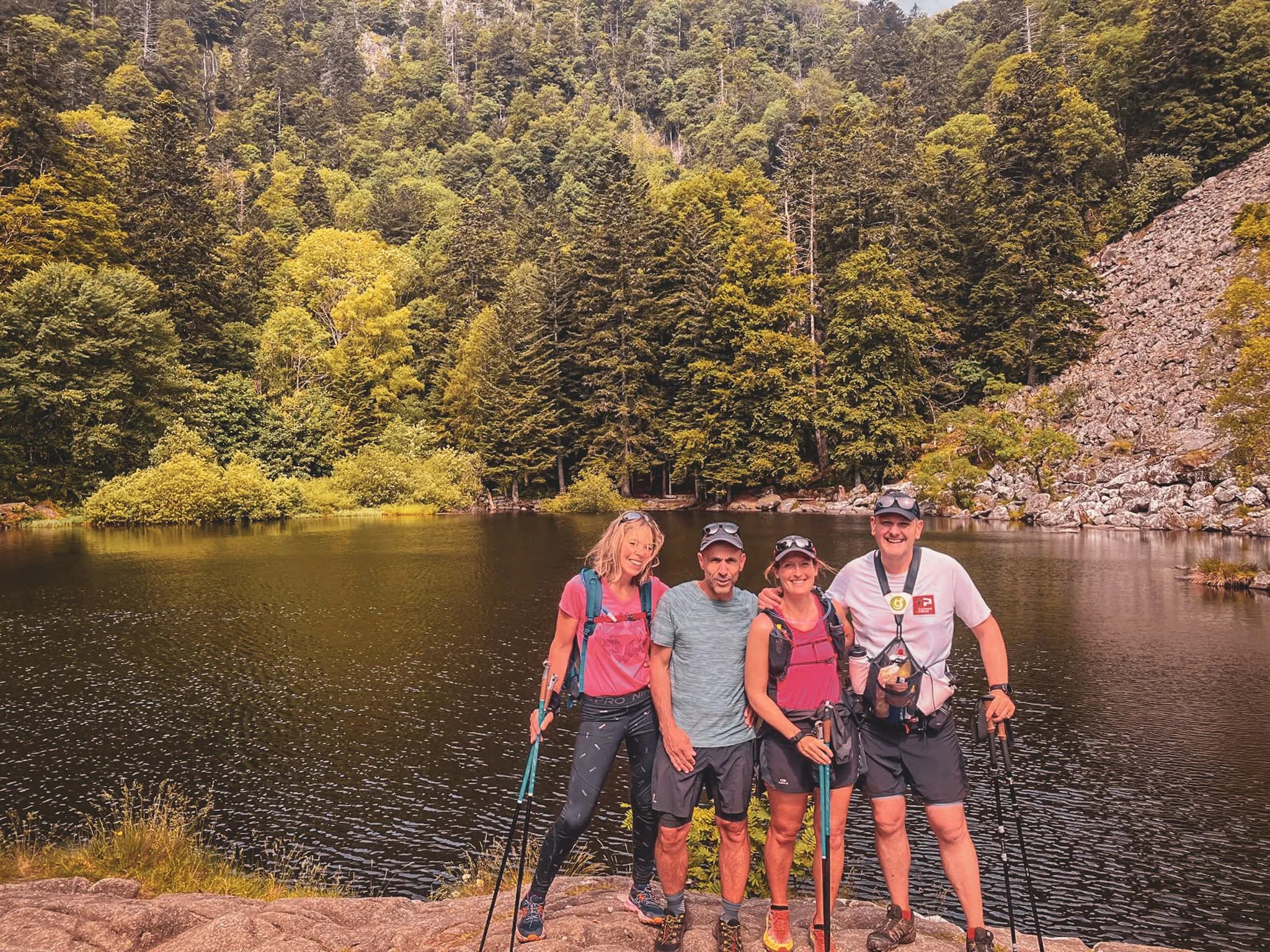 Four smiling hikers beside a lake, surrounded by lush countryside in the Vosges.