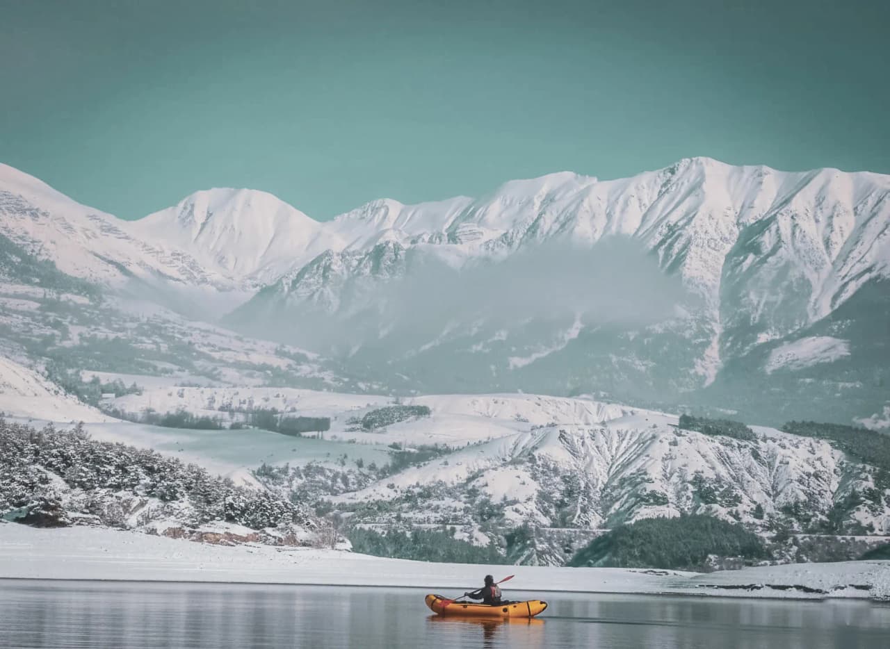 Un aventurier en kayak orange sur un lac glacé, entouré de montagnes enneigées majestueuses.