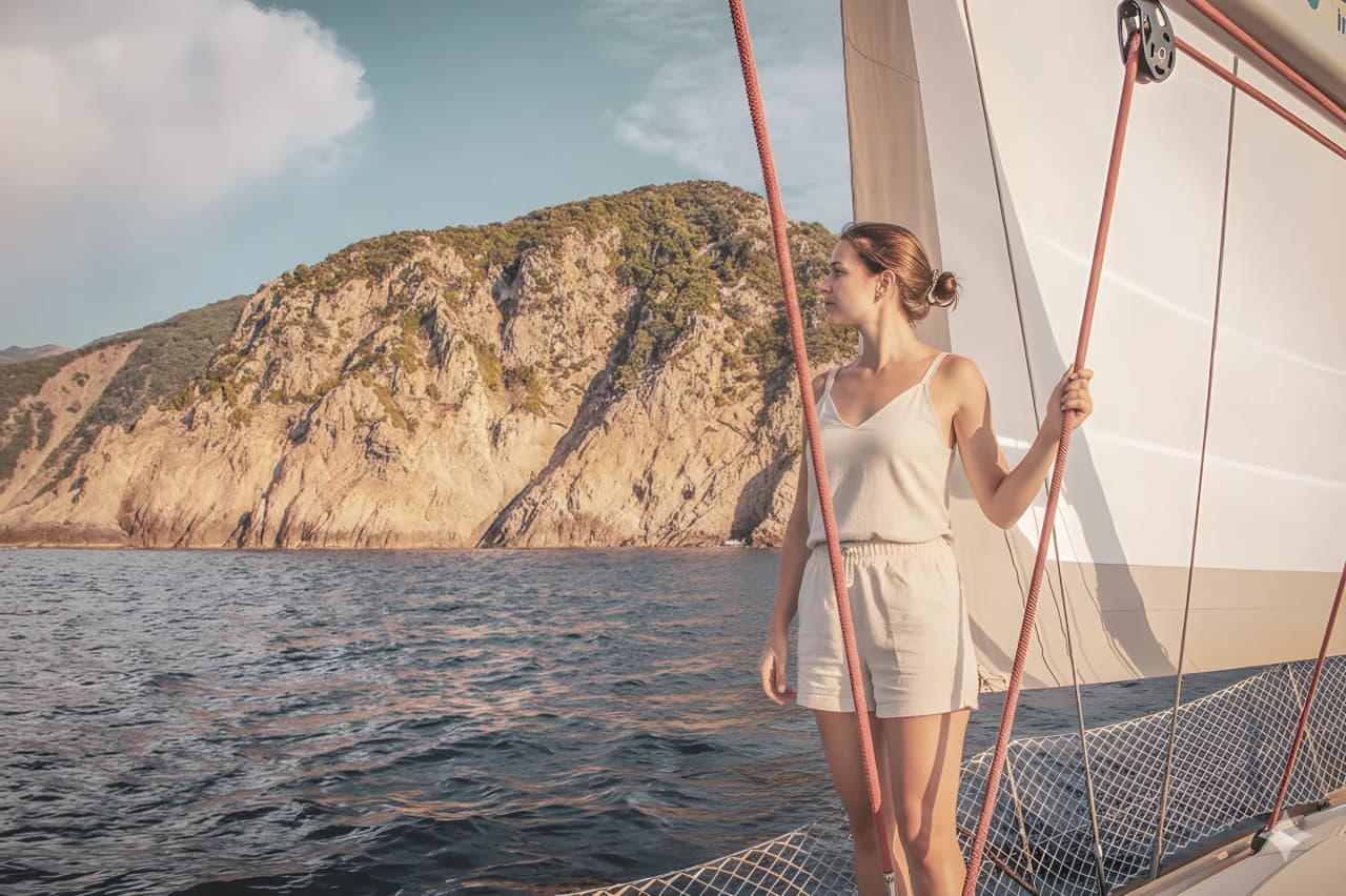 Femme à bord d'un voilier, admirant les falaises ensoleillées de la Corse du Nord.