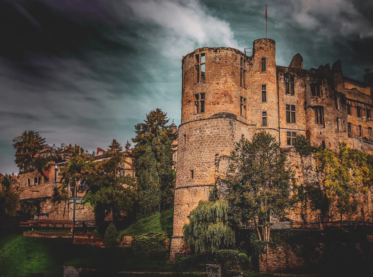 An imposing medieval castle, built of stone, stands proudly on a green hill. The castle walls are adorned with large windows, some with exposed stonework and others more modern. In the