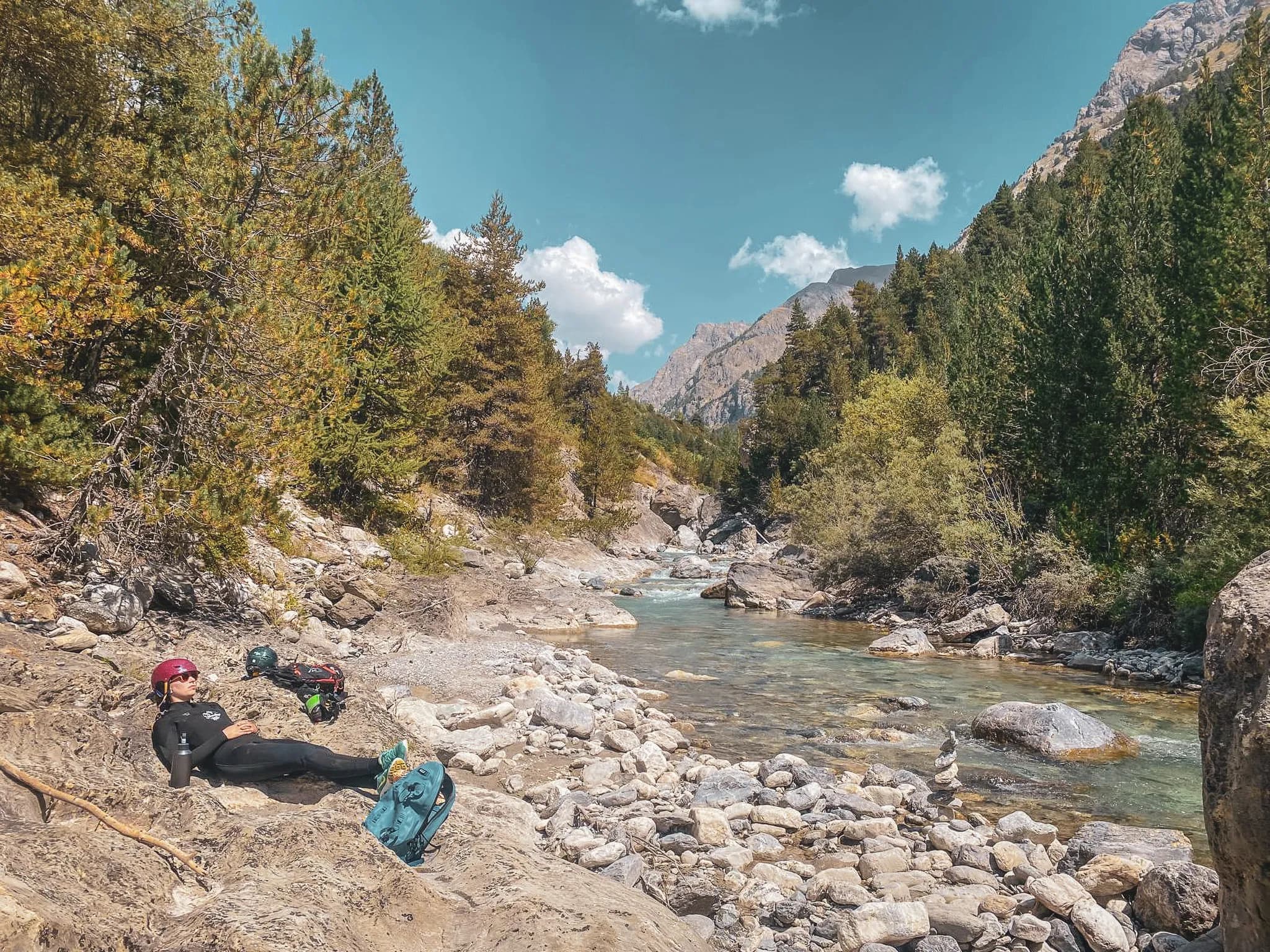 An adventurer in a wetsuit relaxes on the banks of a river surrounded by mountains and forests.