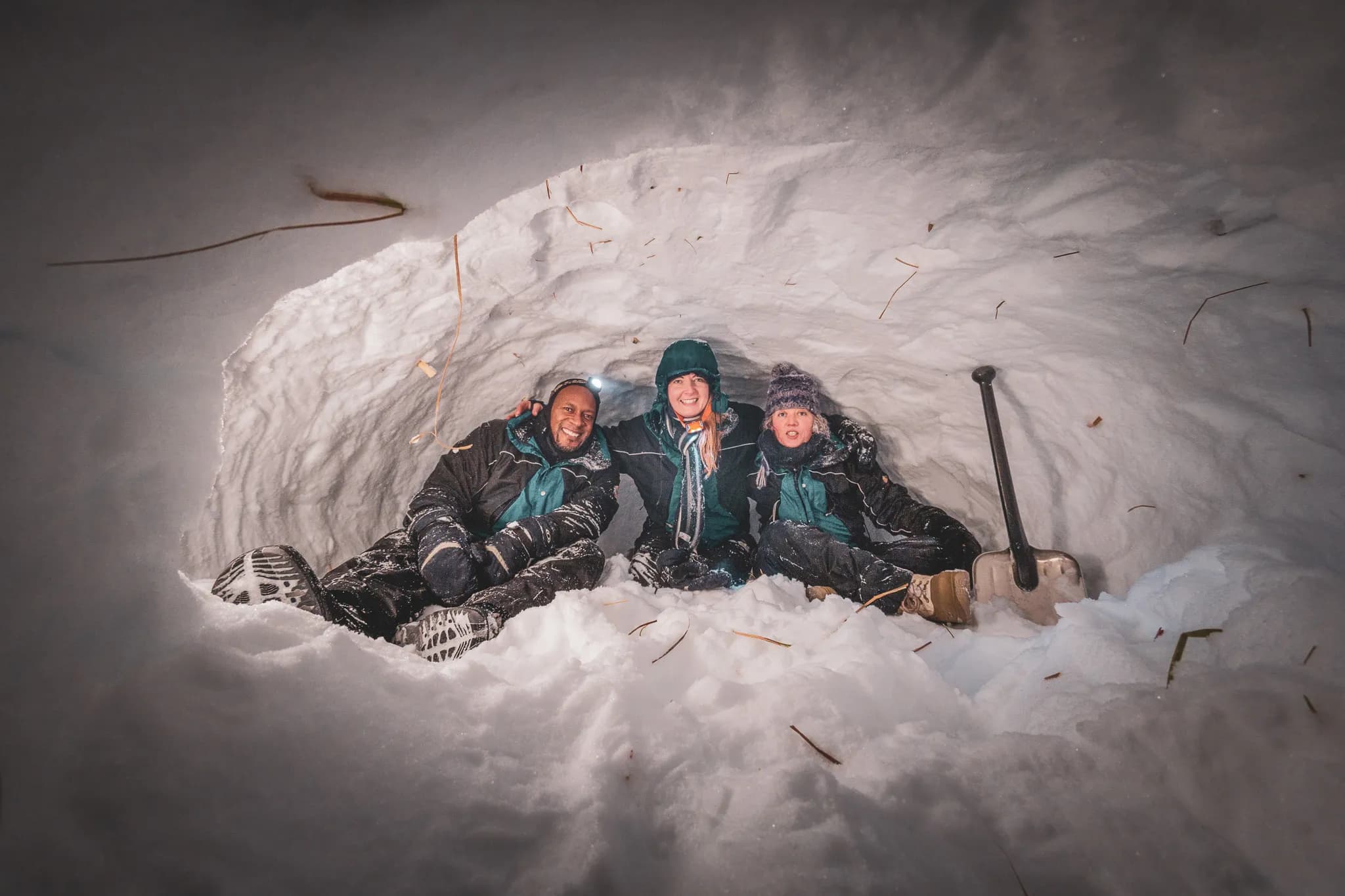 A group of three people sitting inside a snow shelter, similar to an igloo. They are wearing thick winter clothes and look comfortable, despite the cold environment. The soft light illuminates the interior,