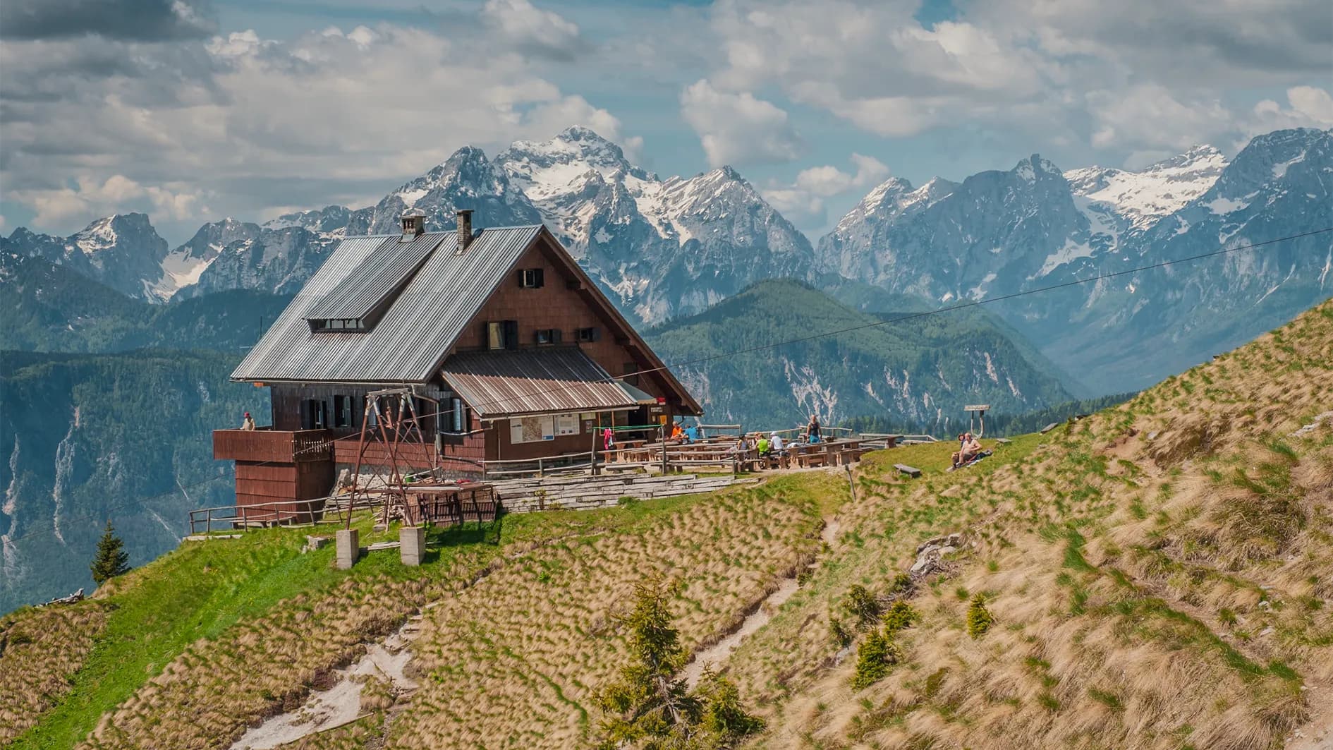 A wooden chalet sits on a green hill, surrounded by majestic mountains, some of which are covered in snow. The sky is partly cloudy, with glimpses of sunshine. By boat, people profit