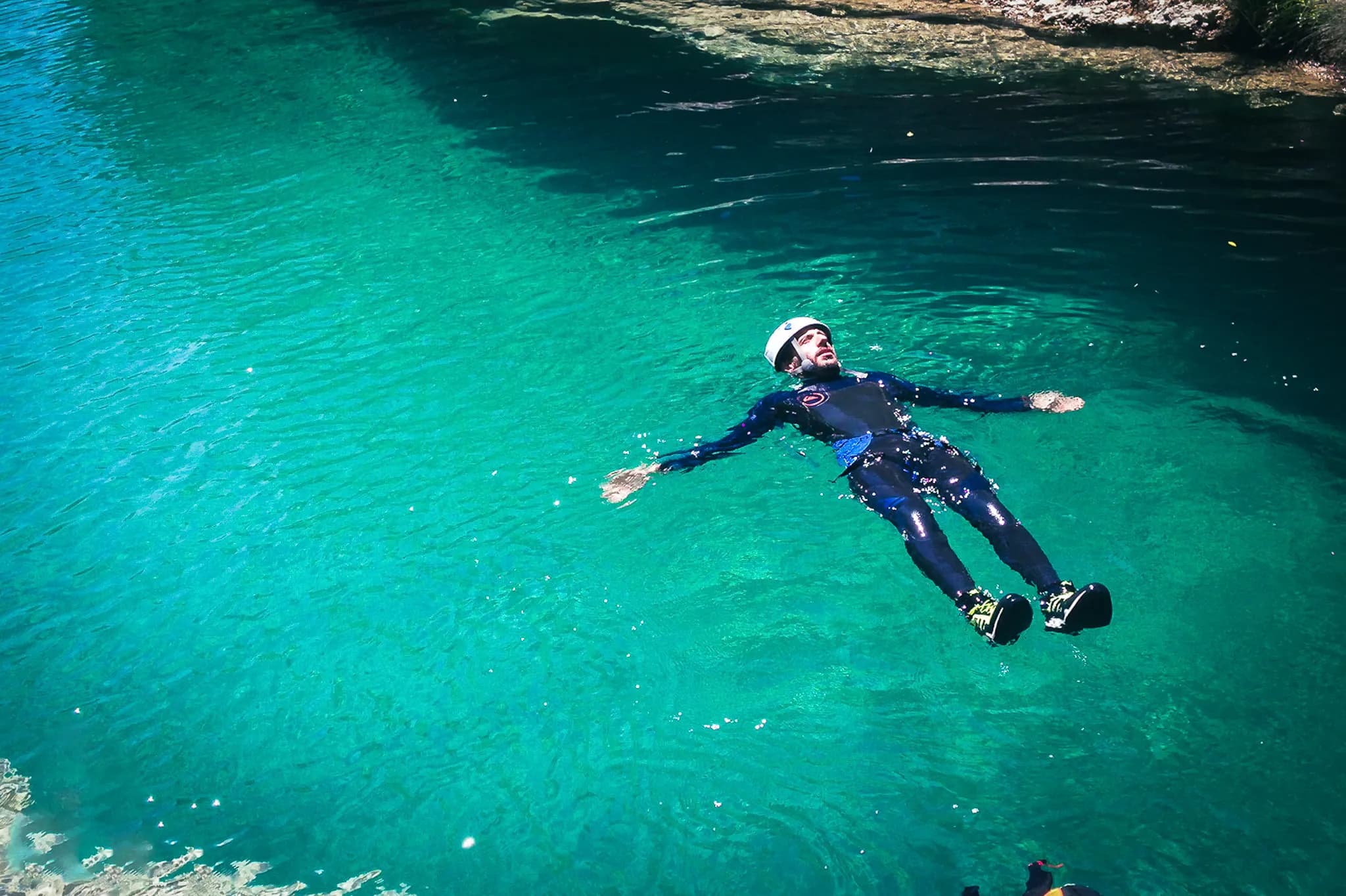 A man floats on crystal-clear, bright blue water in a natural landscape. He is wearing a black wetsuit with blue accents and a helmet, seemingly enjoying a sunny day. The