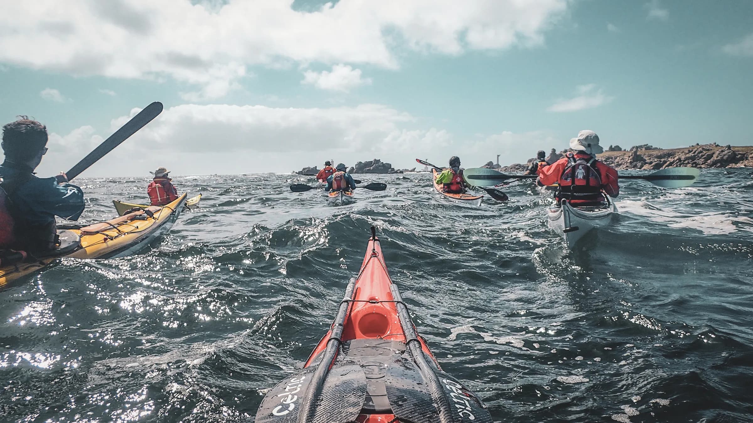 A group of kayakers sailing on white water, with islands in the background and a clear sky.