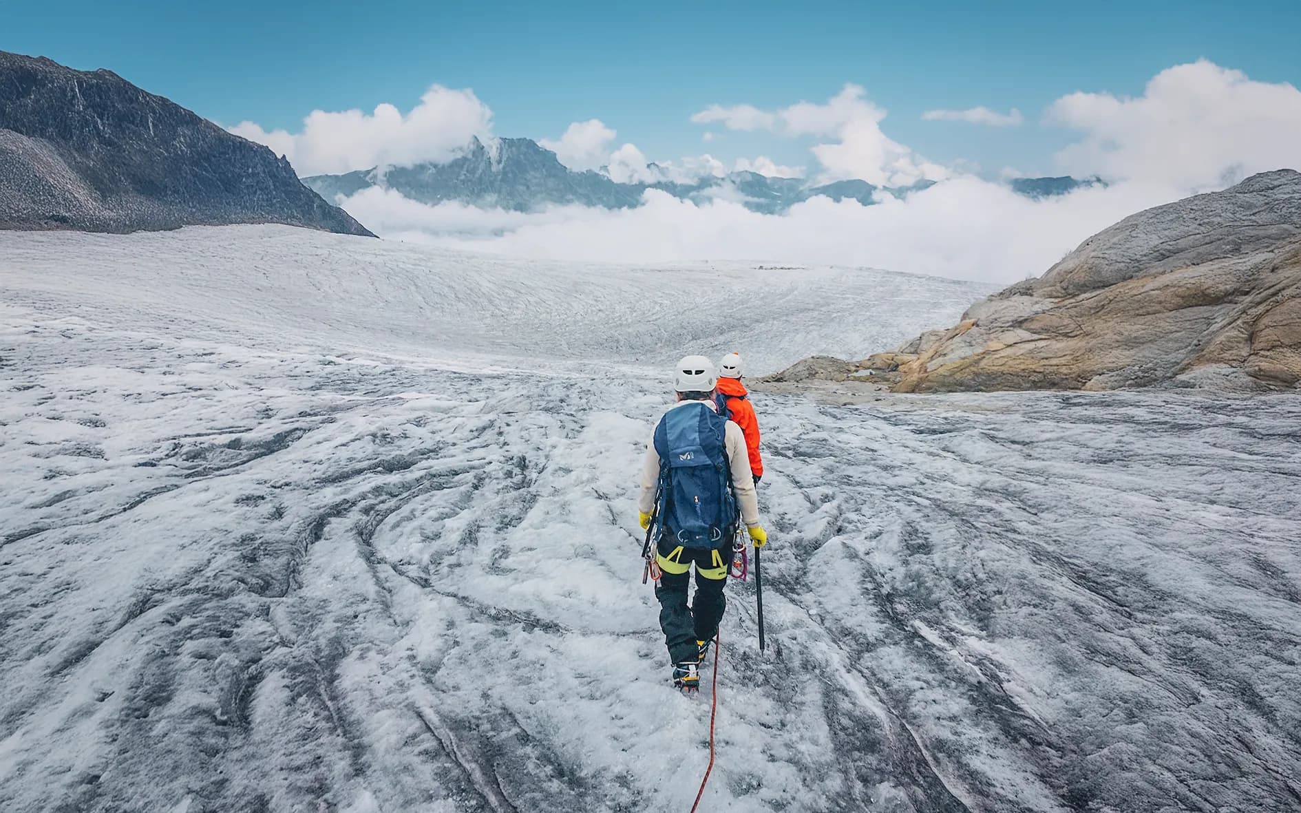 An expedition on a glacier is featured, with two climbers making their way across an icy surface in shades of grey and white. The surrounding landscape is mountainous, with summits slightly misted by