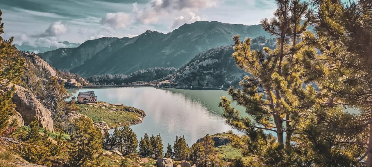 A peaceful lake surrounded by mountains, with a waterside Mountain hut under a bright sky.