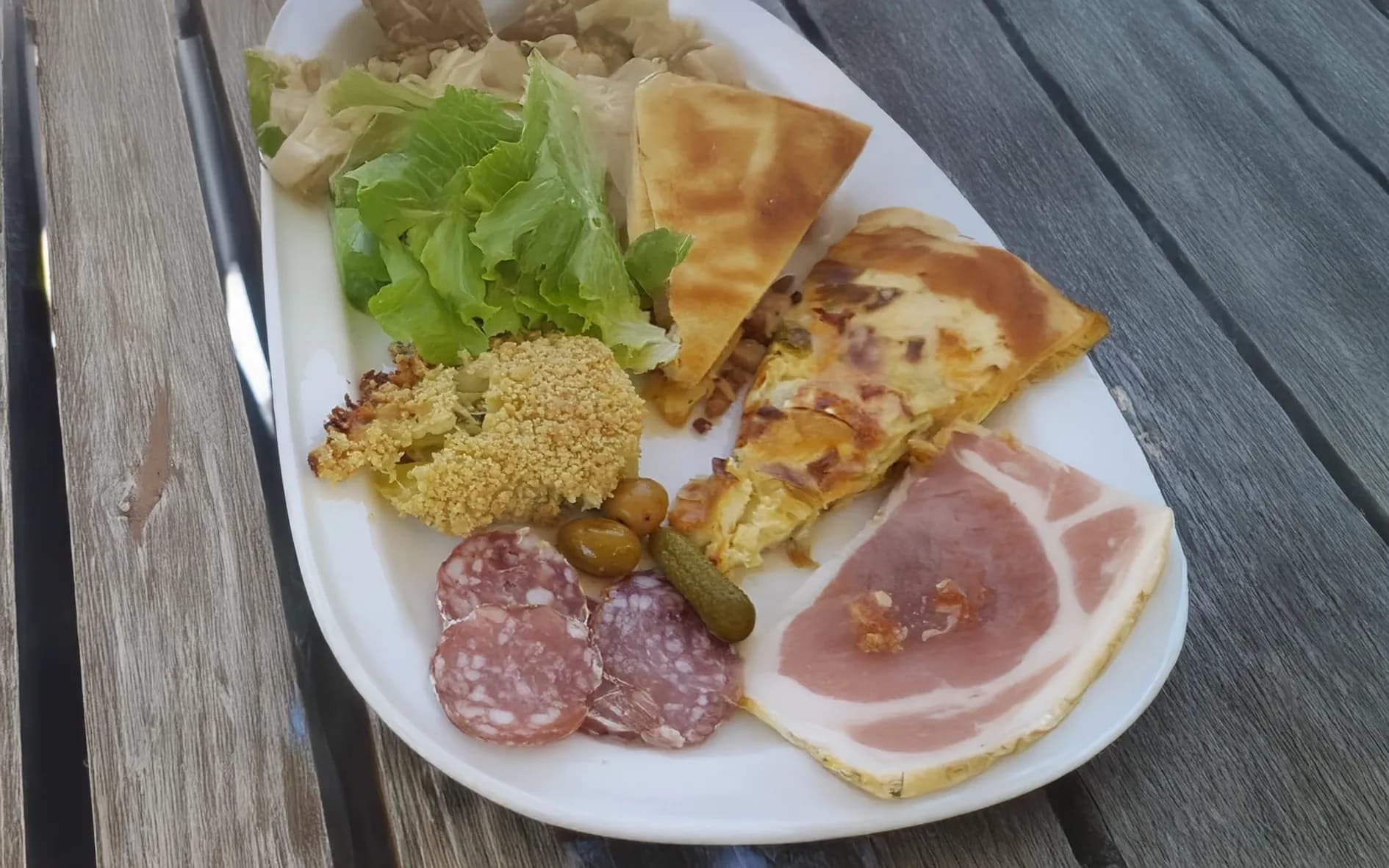 A plate of cold meats and fresh vegetables is presented. There's a salad of lettuce leaves and a piece of roasted cauliflower. There are also slices of charcuterie,