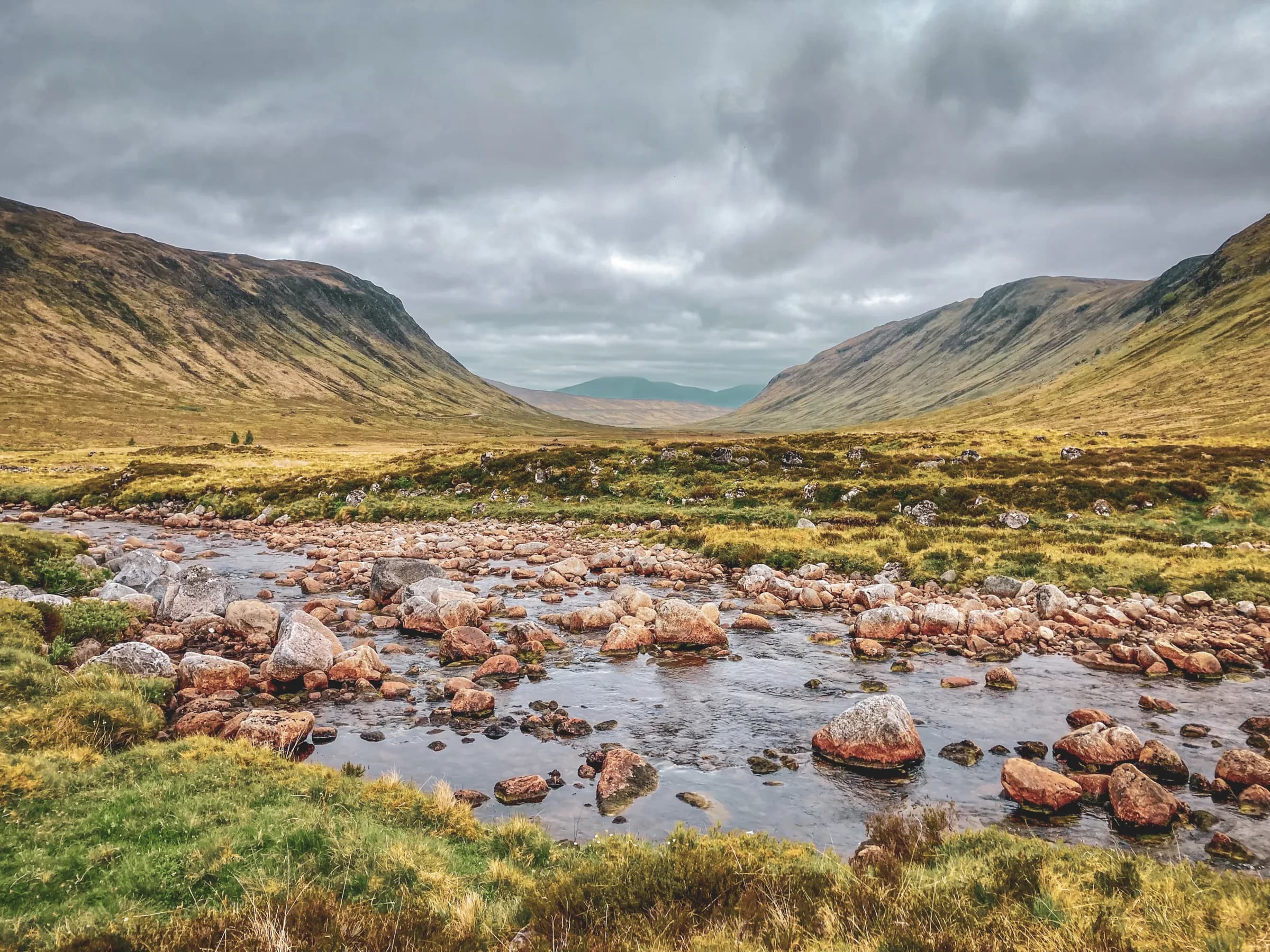 A vast Scottish valley with a stream winding between rocks, under a cloudy sky.