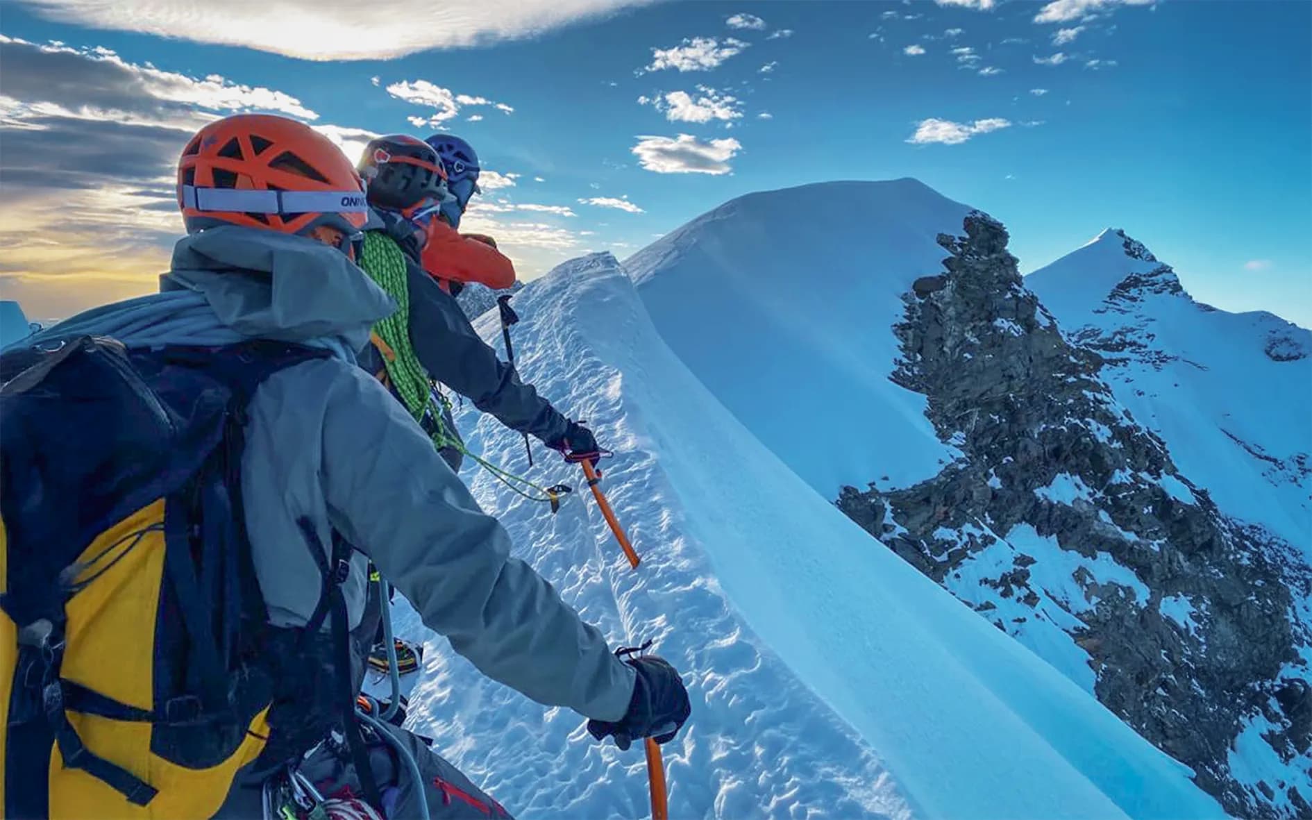 A group of climbers stand on a snow-covered ridge, observing the surrounding landscape. They are wearing orange and blue safety helmets and climbing gear suitable for the cold. Each of them is