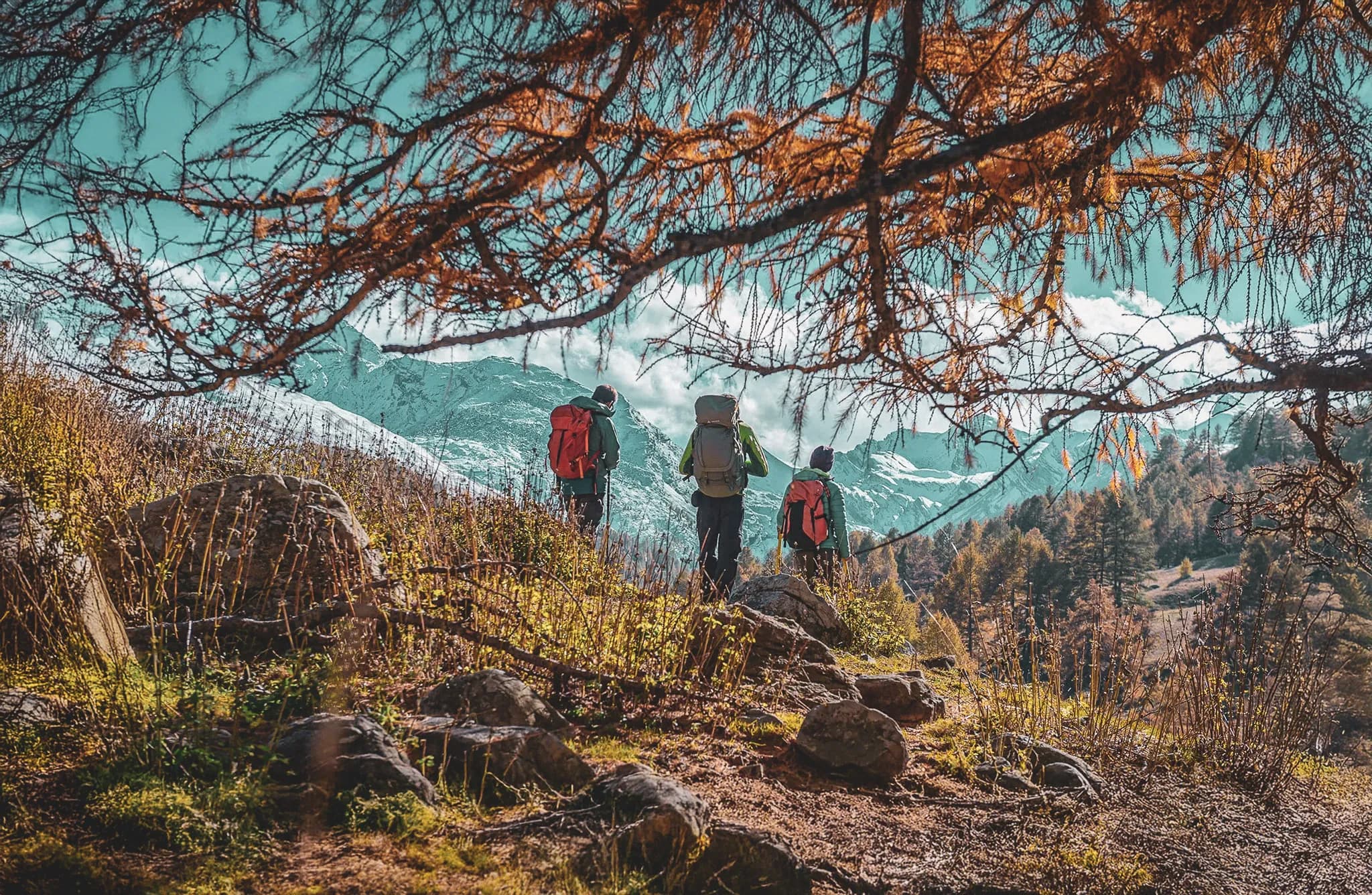 A group of hikers stand on a mountain path, admiring the landscape. They are carrying rucksacks and look ready to explore. In the background, majestic mountains rise up, partially covered by the snow.
