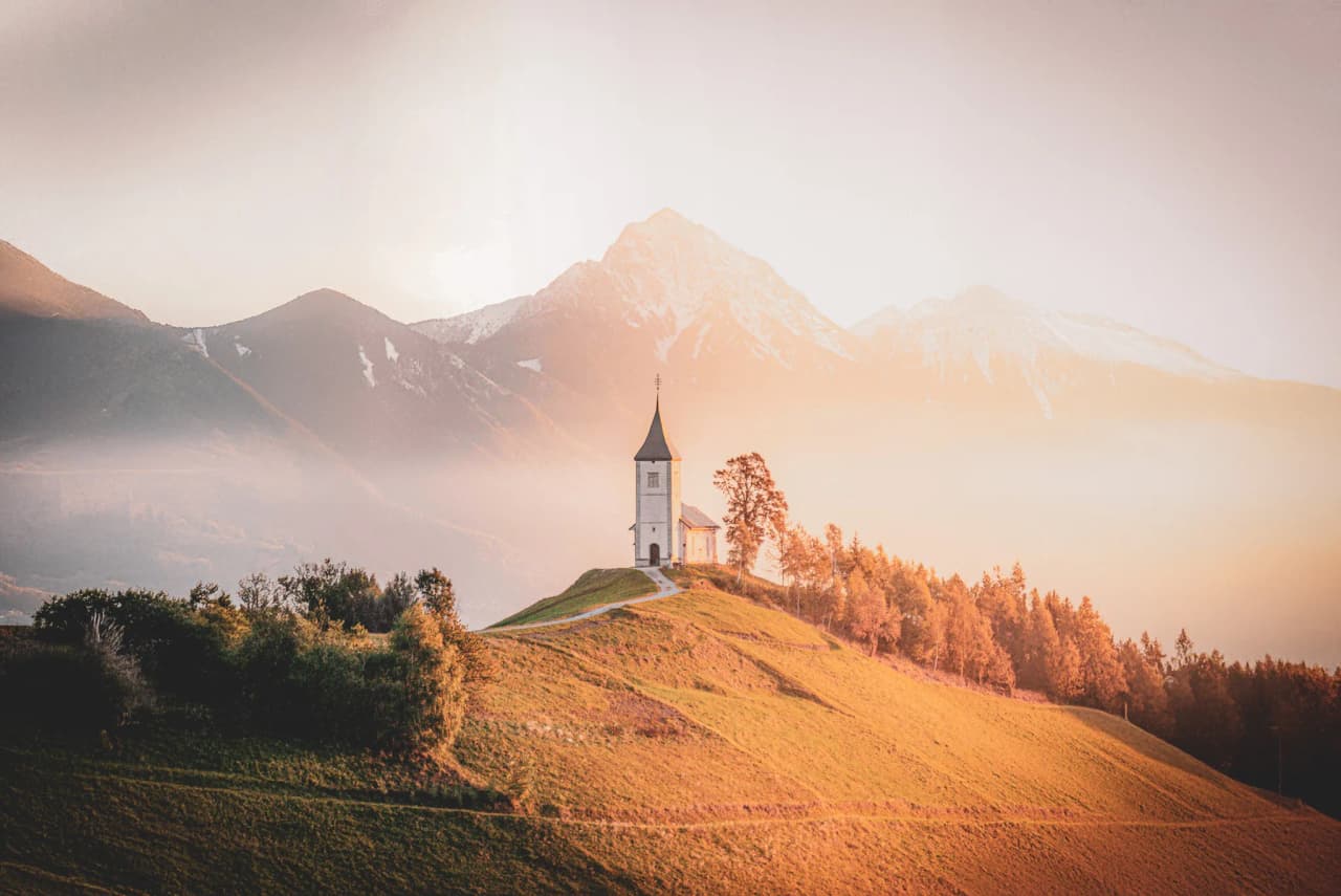 A kayaker navigates a river surrounded by green mountains and large rocks. The landscape is lush, with leafy trees lining the banks. The sky is partly cloudy, adding