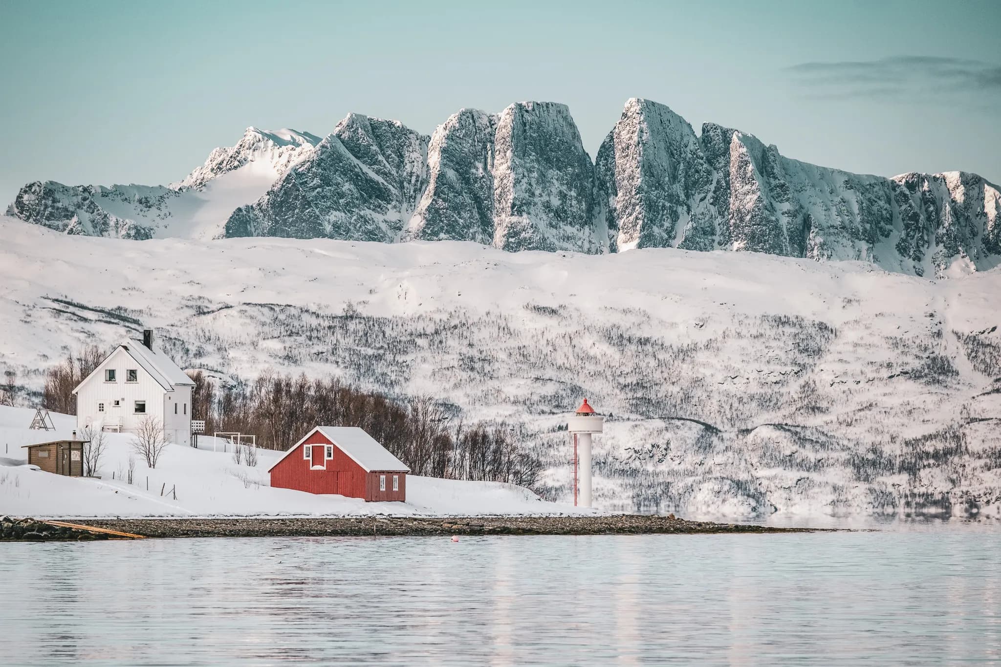 A winter landscape with snow-capped mountains in the background, overlooking a calm, reflective stretch of water. On the left, a white house is surrounded by snow, while on the right is a small red building.