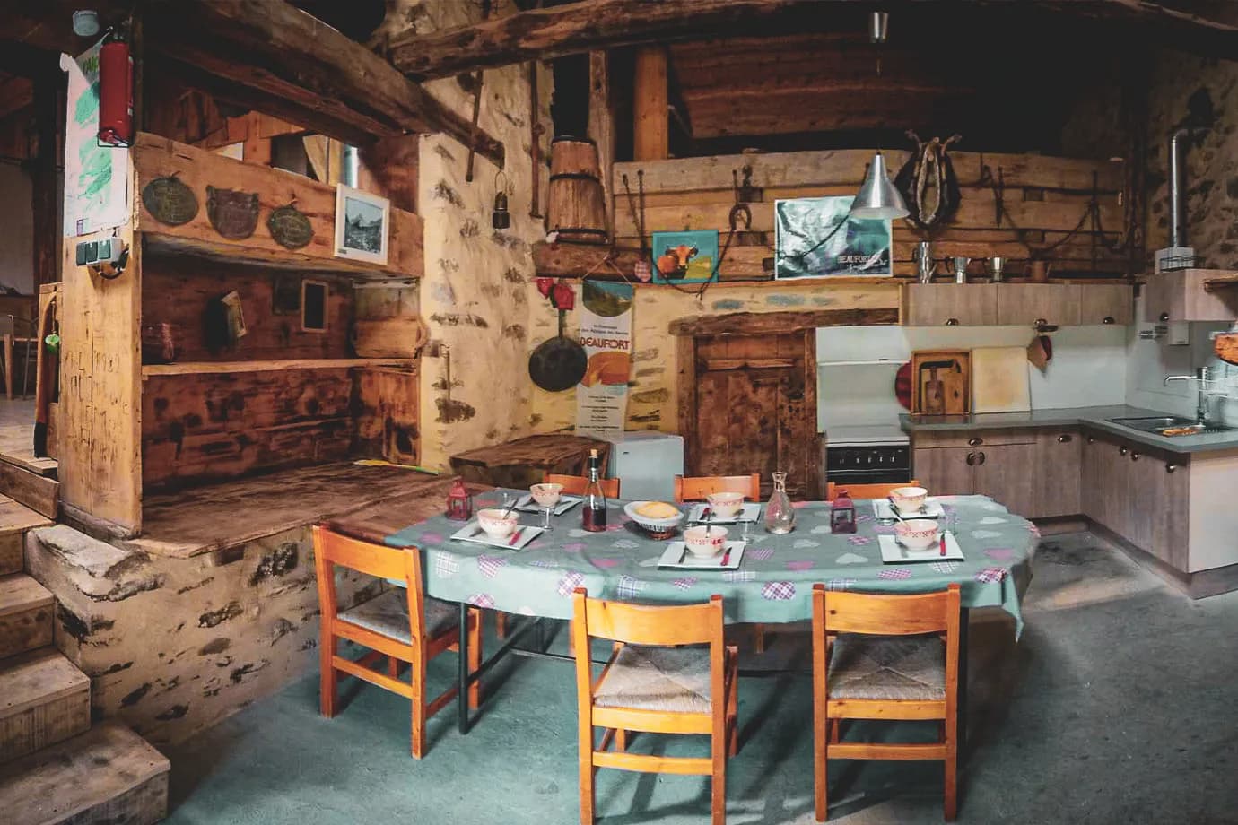 A rustic kitchen with exposed stone walls and wooden beams on the ceiling. A large round table is decorated with a colourful patterned tablecloth, surrounded by wooden chairs. On the table are a number of