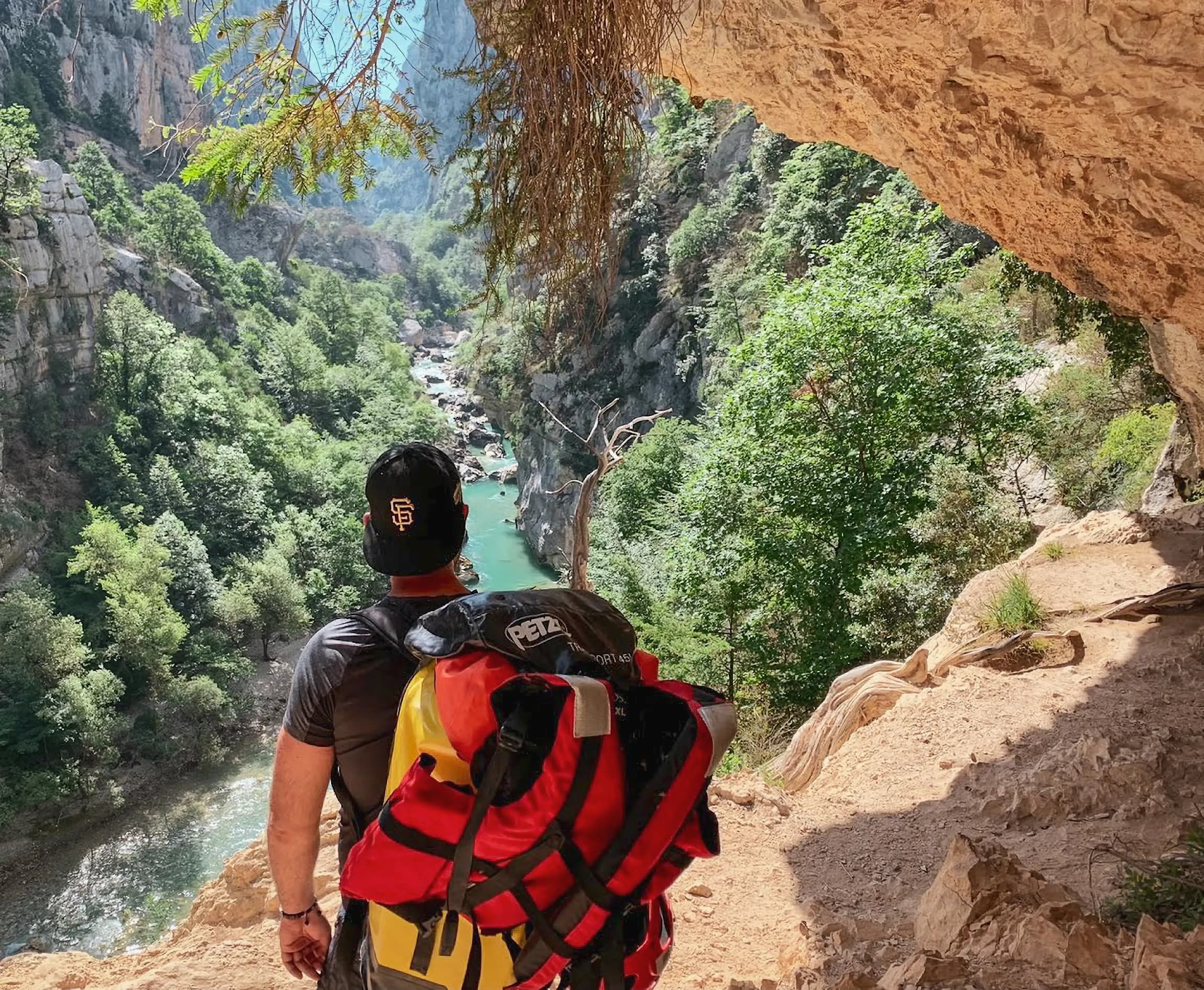 A contemplative hiker stands on a steep path overlooking a green valley where a stream winds its way through rocks and dense vegetation. He is carrying a large rucksack with