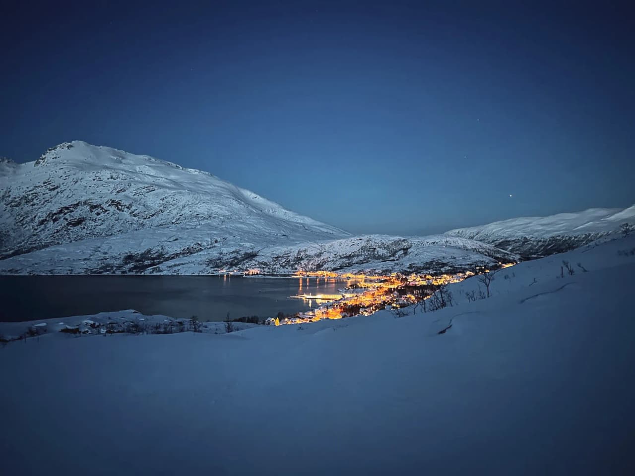 Nachtelijk uitzicht op besneeuwde bergen en een verlicht dorp aan het water in Tromsø.