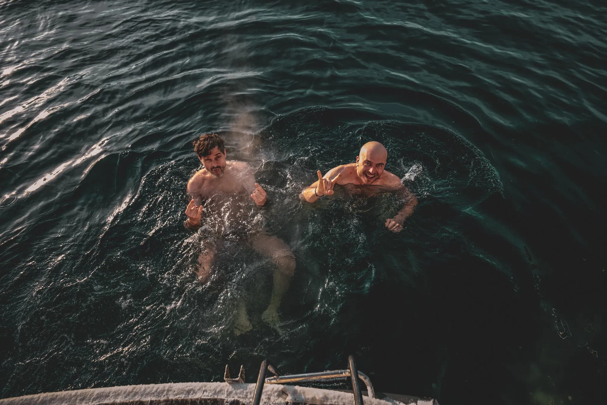 Deux hommes se trouvent dans une eau sombre, en train de nager. L'un d'eux, avec les cheveux bruns et une moustache, fait un geste avec les mains, semblant s'amuser. L'autre, chauve,
