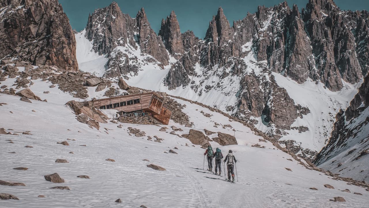 Trois randonneurs marchent vers un refuge pittoresque, entouré de majestueuses montagnes enneigées.