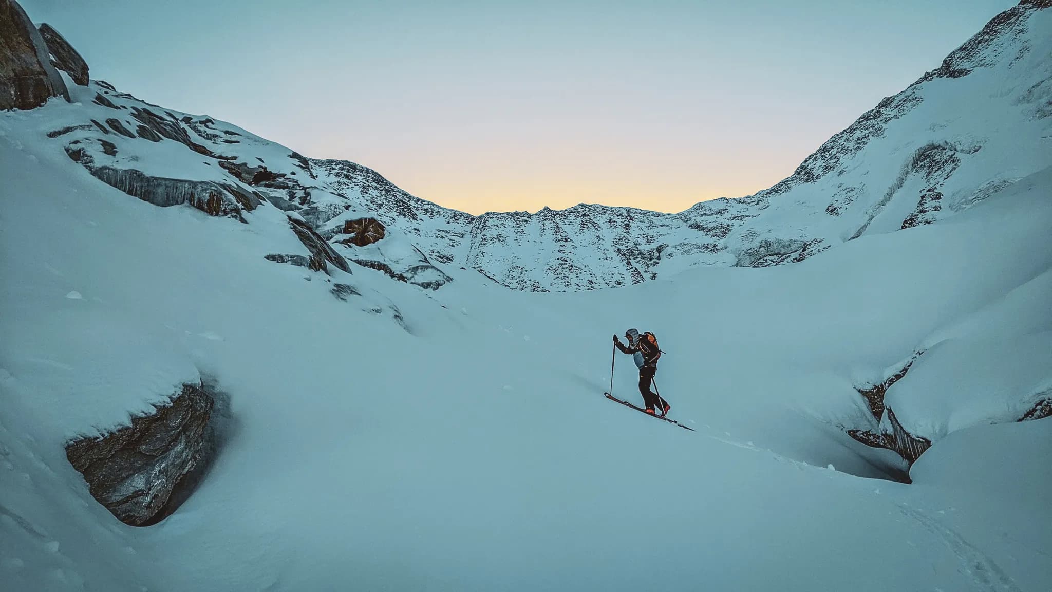Un skieur progressant dans un paysage de montagne enneigé, entouré de flancs rocheux et de neiges compacts. La lumière du crépuscule illumine l'horizon, créant un contraste avec les sommets majest