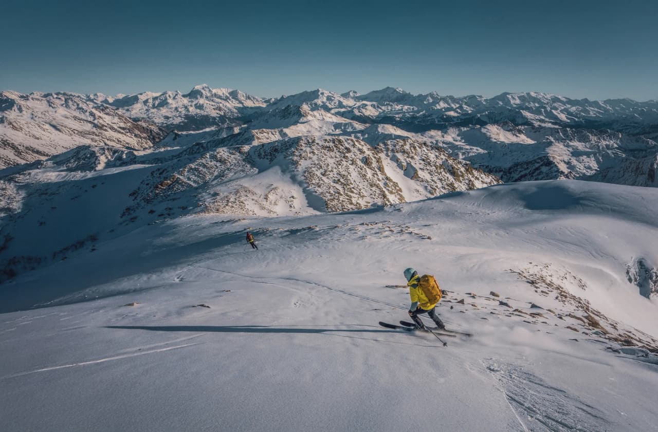 A snowy mountain landscape with skiers hurtling down the slopes. The scene features majestic Alpine peaks in the background, under a brilliant blue sky. Ski tracks mark the