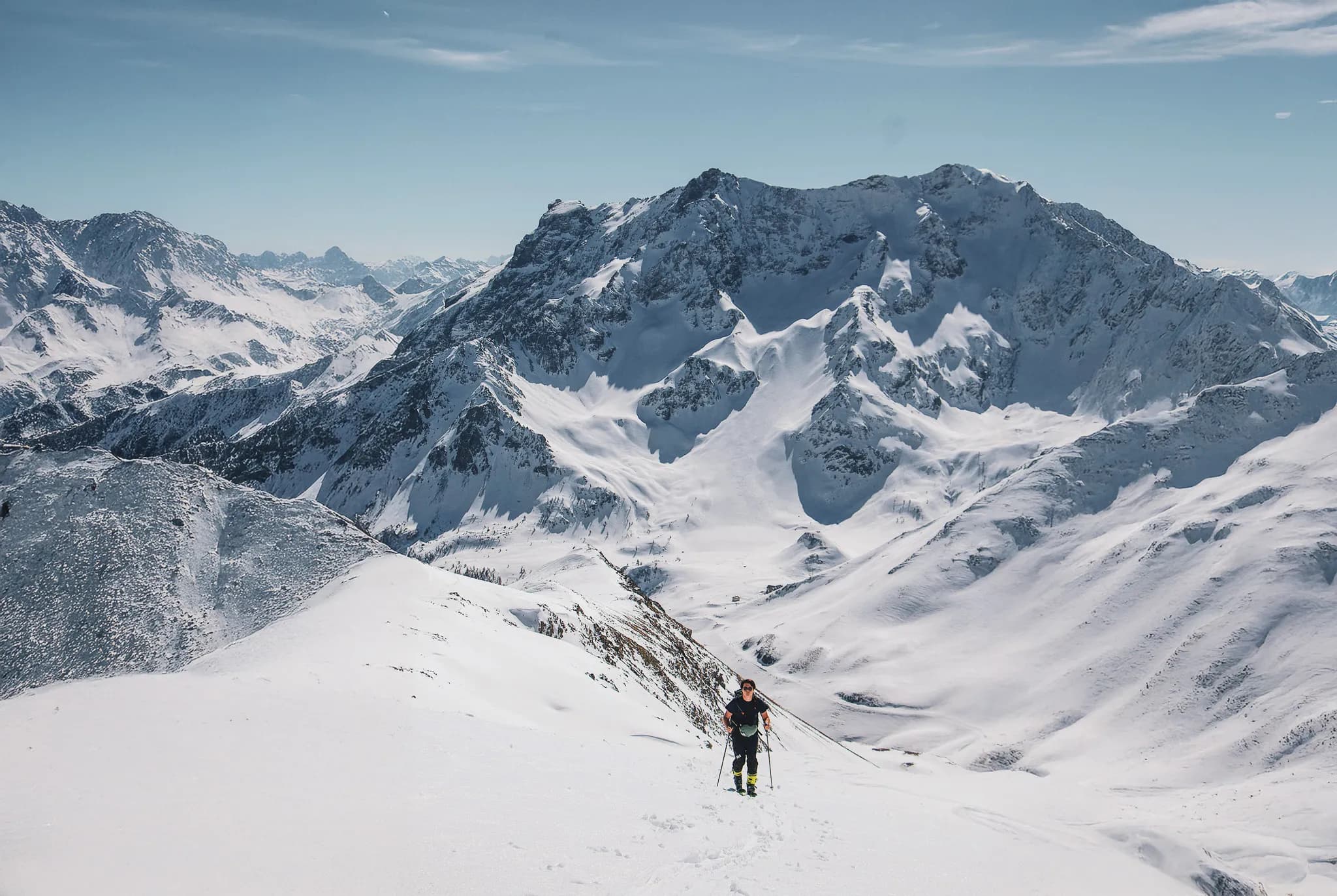 Un aventurier en pleine ascension dans un paysage montagneux majestueux, entouré de vastes étendues de neige. Les montagnes, couvertes de neige éclatante, se dressent sous un ciel clair avec quelques nuages, cré