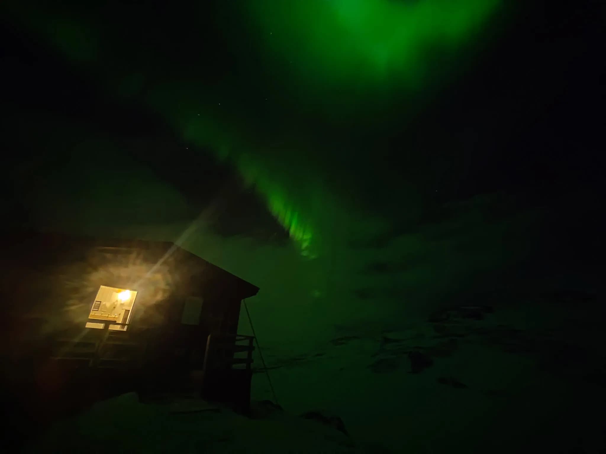 An STF wooden chalet stands in a night landscape, illuminated by the glow of yellow light from a window. Above, the northern lights illuminate the sky with brilliant green hues.