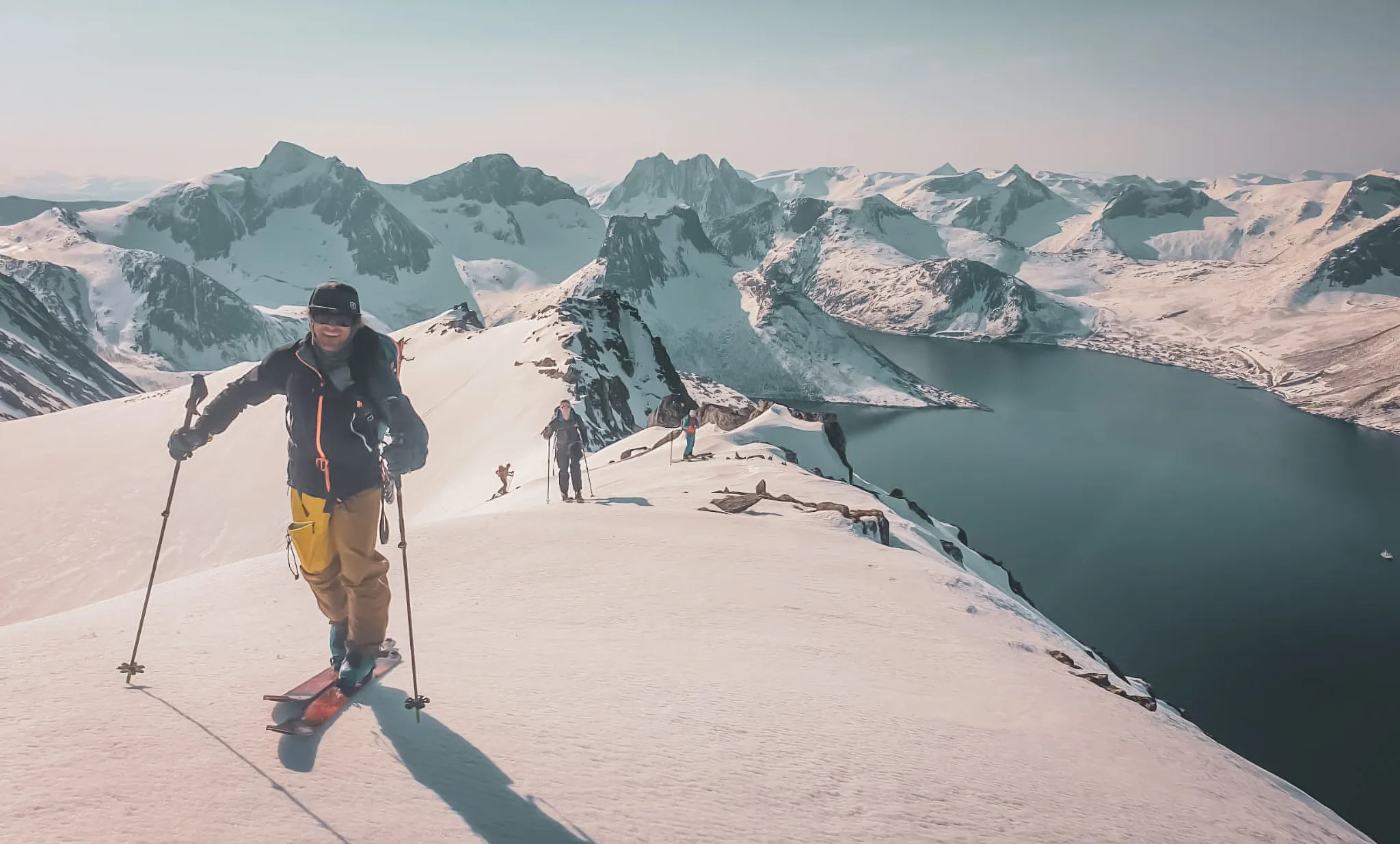 Een lachende man skiet over een besneeuwde bergkam, omringd door spectaculaire besneeuwde bergen en een meer beneden. Silhouetten van andere skiërs doemen op in de achtergrond en voegen een avontuurlijke dimensie toe.