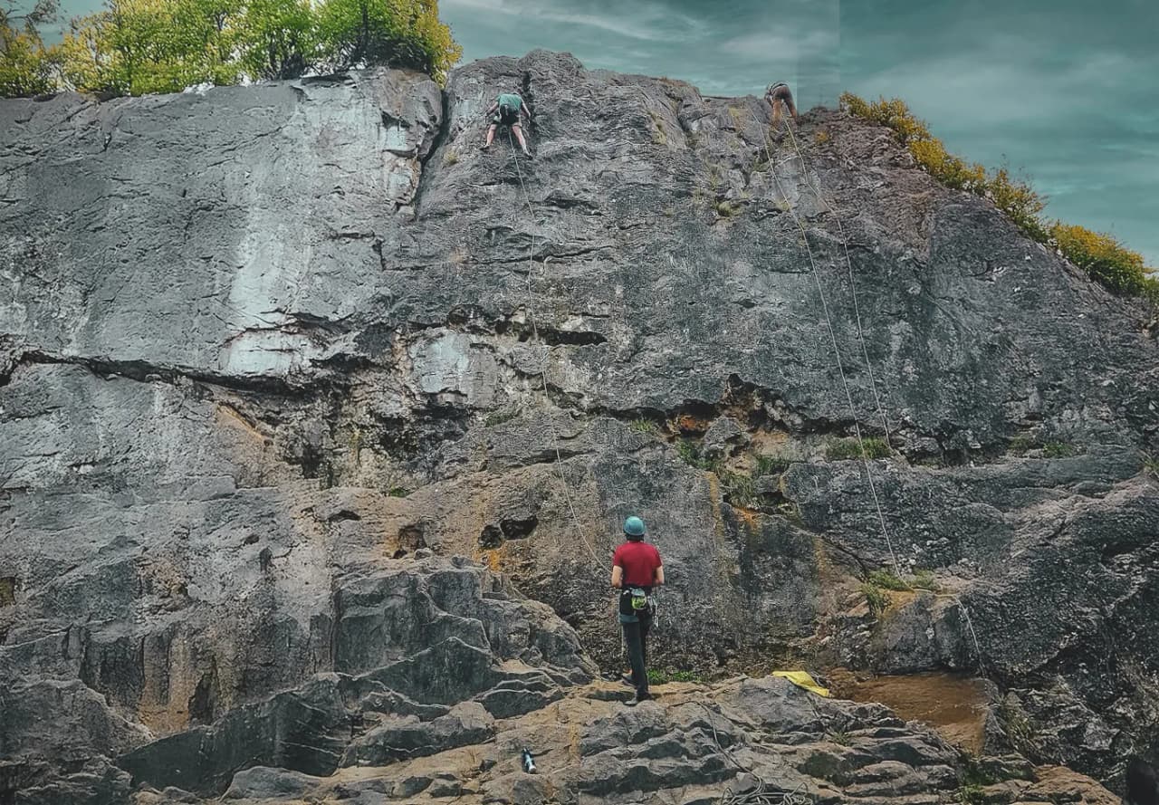 A climber is scaling a rocky cliff, while another climber is already at the top, against a backdrop of cloudy skies. Down below, someone is observing the scene, wearing a helmet and climbing harness.