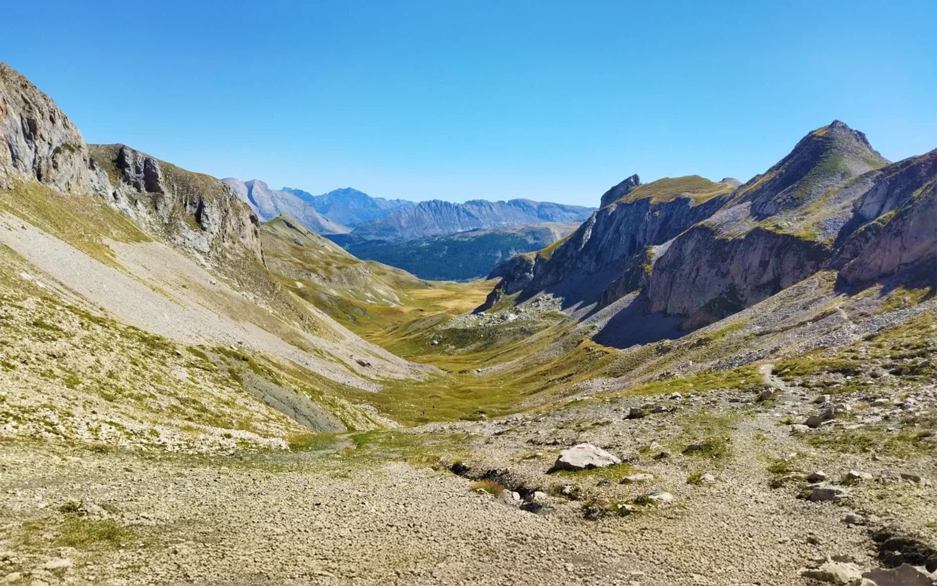 A vast mountain landscape, with craggy, rocky peaks rising up against a clear blue sky. The valley below is blanketed in greenery and golden meadows, while rocks and