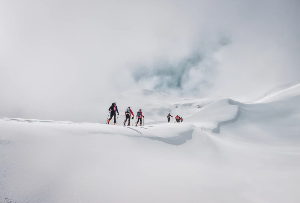 A group of mountain skiers move across virgin snow, surrounded by Alpine scenery in a misty atmosphere. The skiers, dressed in colourful clothing and carrying ski poles, cross the