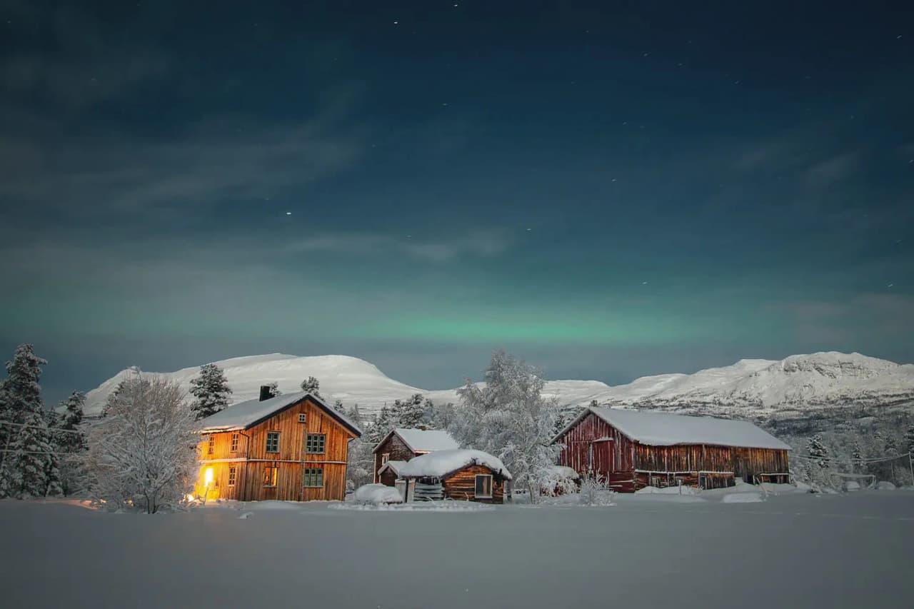 A peaceful winter landscape with several wooden chalets surrounded by snow. One of the cottages, with its illuminated windows, looks cosy in the dark. In the background, hills rec