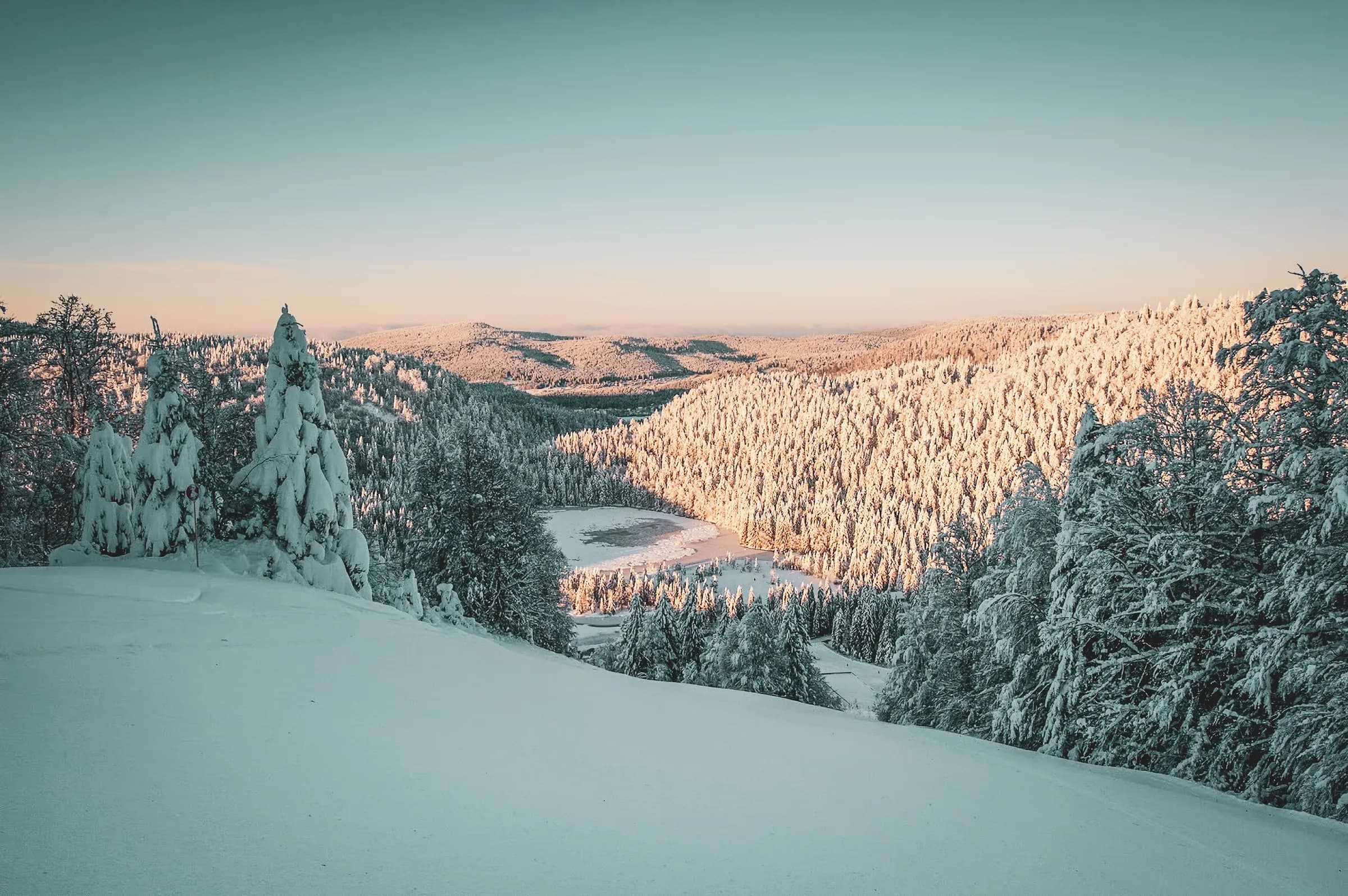 Een betoverend winterlandschap met besneeuwde heuvels. Majestueuze besneeuwde bomen strekken zich uit zover het oog reikt en vormen een dicht bos. In het midden, een gedeeltelijk bevroren meer