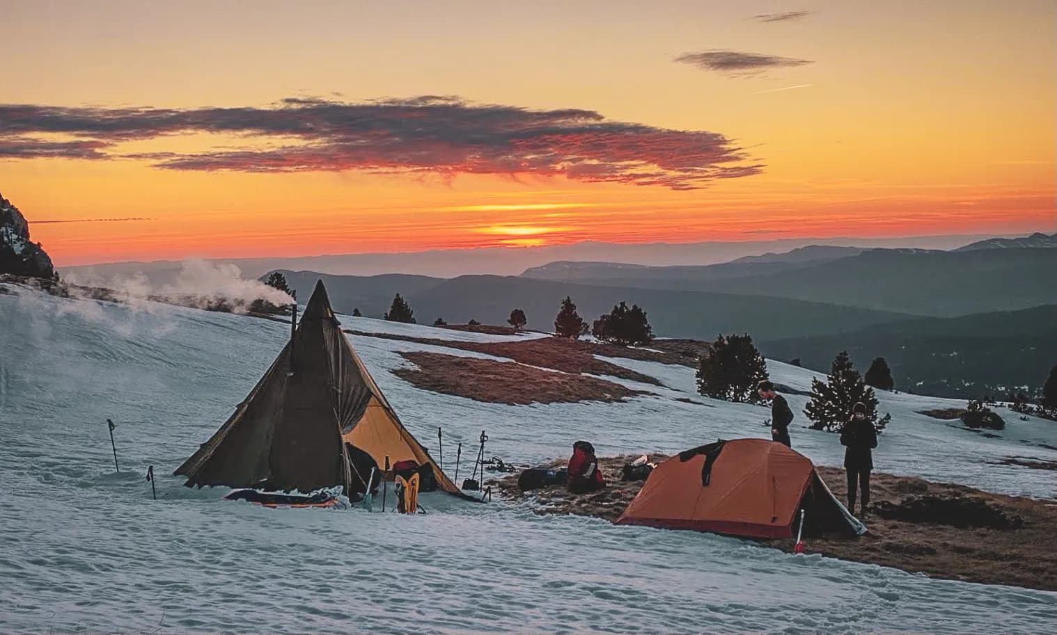 A mountain landscape at dusk, with tents pitched on snow-covered ground, surrounded by conifers. The sky is tinged with shades of orange and pink, illuminated by a sunset. The tent