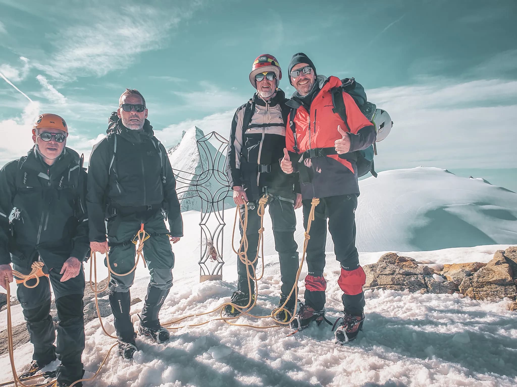 Four smiling climbers pose on a glacier, surrounded by majestic peaks under a blue sky.