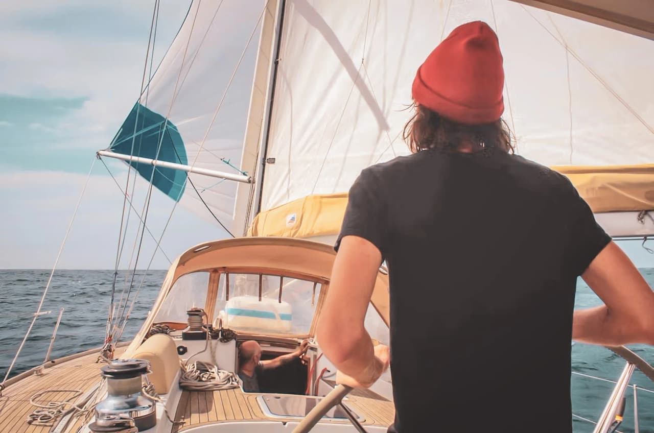 Un marin à la barre d’un voilier, aux eaux turquoises, sous un ciel ensoleillé. Aventure bretonne!