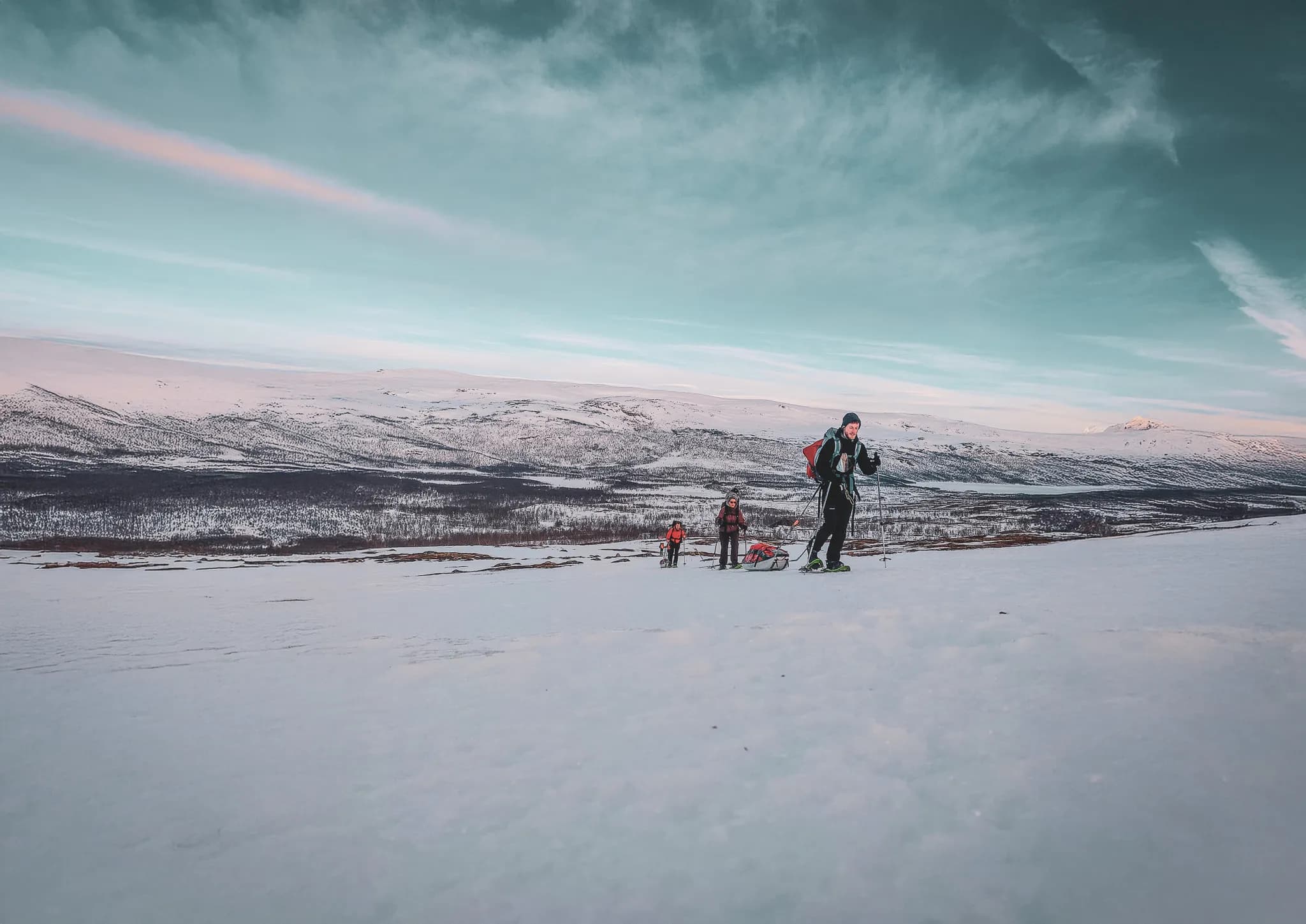 A group of people hiking on snow-covered ground, with mountains in the background. One of the people in front is smiling and using hiking poles, while the others follow, carrying packs.