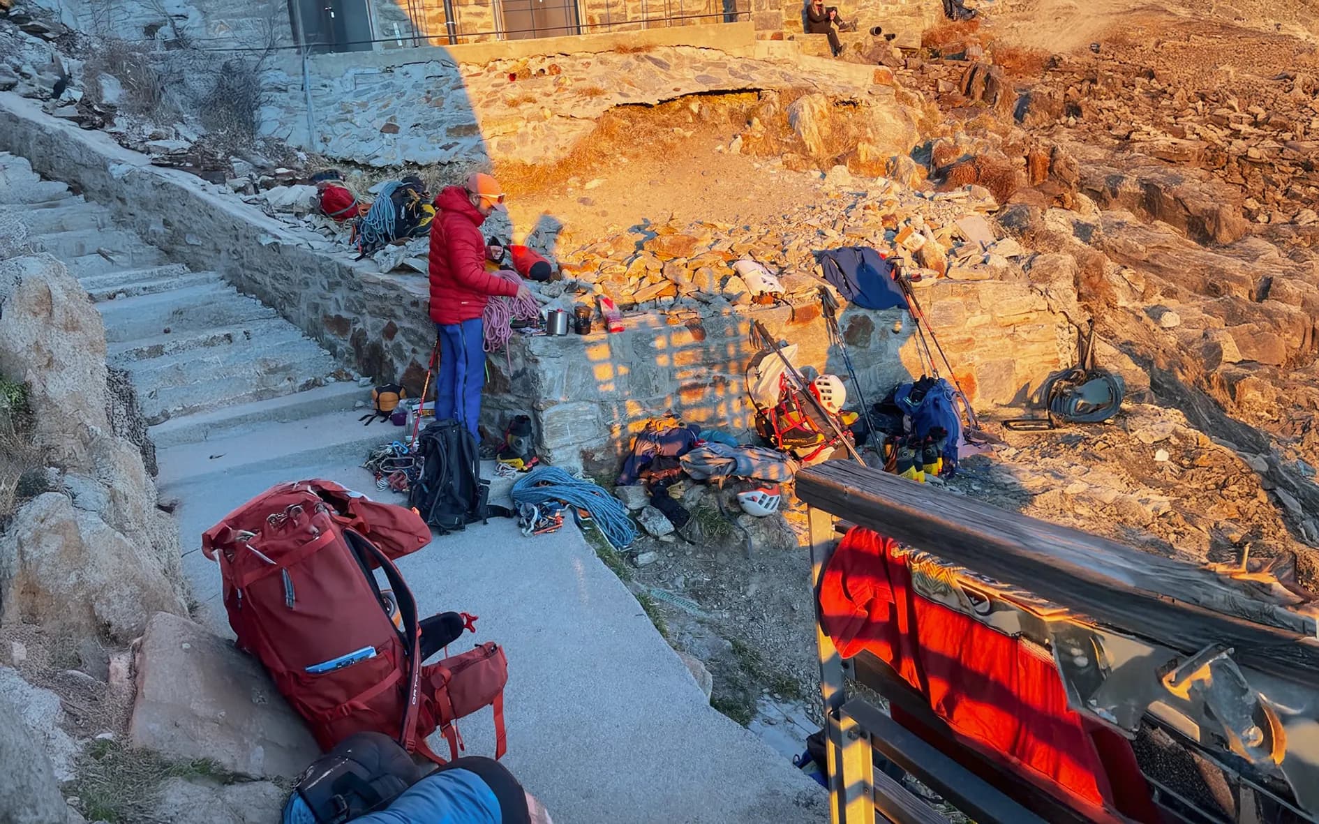 A group of people prepare to climb, situated on a stone platform with steps leading down to rocky terrain. Ropes and climbing equipment are scattered around, while one person in