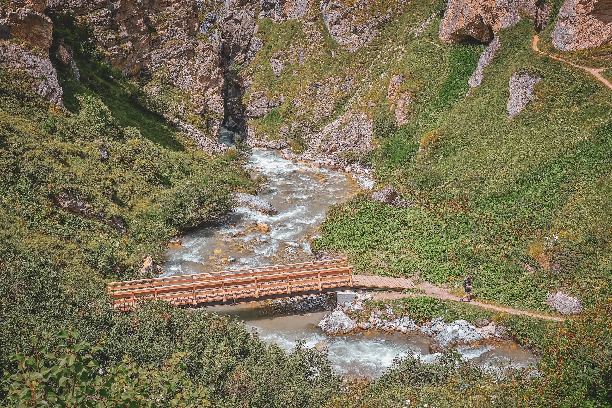 Un panorama montagneux spectaculaire dévoile des sommets majestueux aux arêtes découpées. À l'avant-plan, un sentier sinueux traverse des pentes verdoyantes parsemées de rochers. À gauche, quelques chalets en