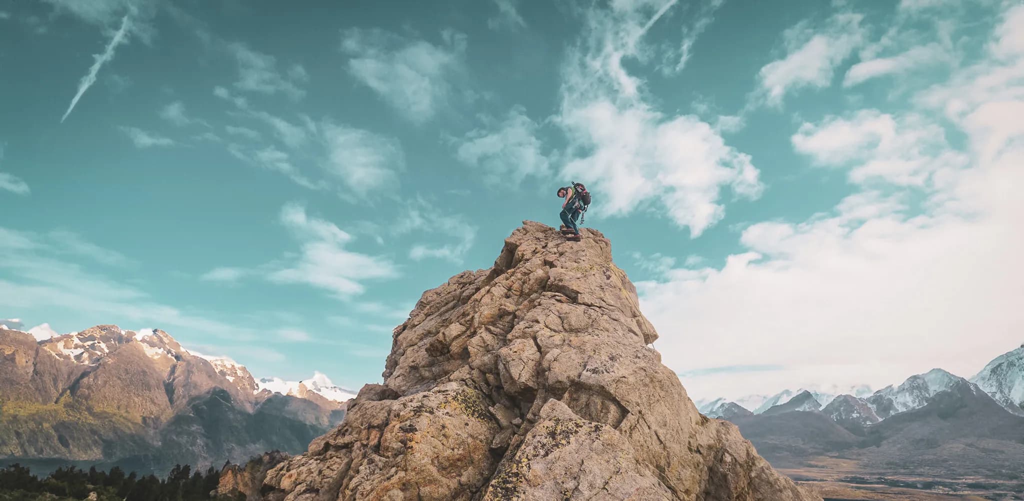 A climber stands at the top of an impressive crag, overlooking a spectacular mountain landscape. The sky is a clear blue, dotted with light white clouds and streaks left by