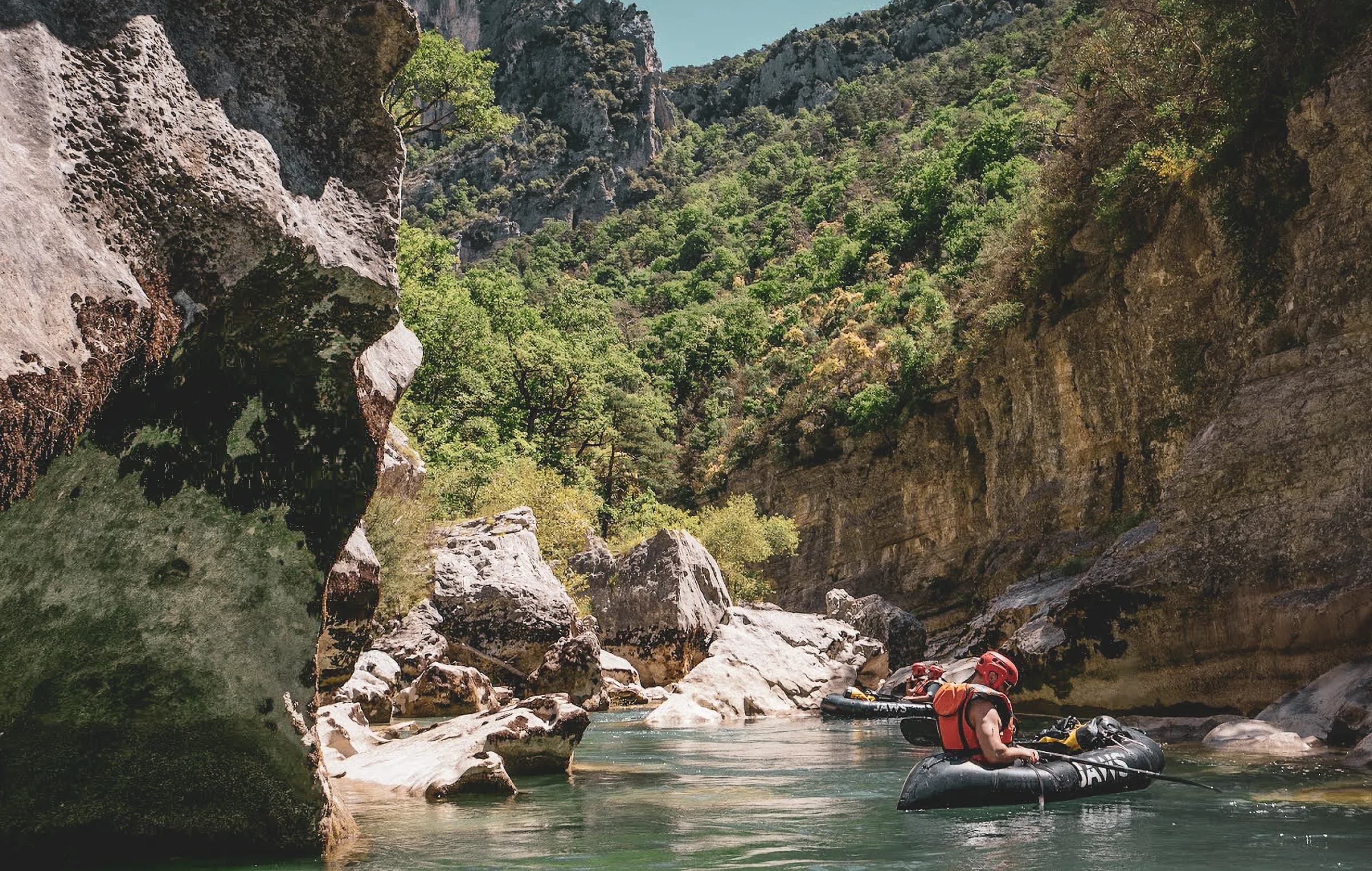 An impressive natural landscape with high cliffs bordered by lush vegetation. A calm river meanders through large rocks, and two people in kayaks float quietly, wearing safety helmets. Visit