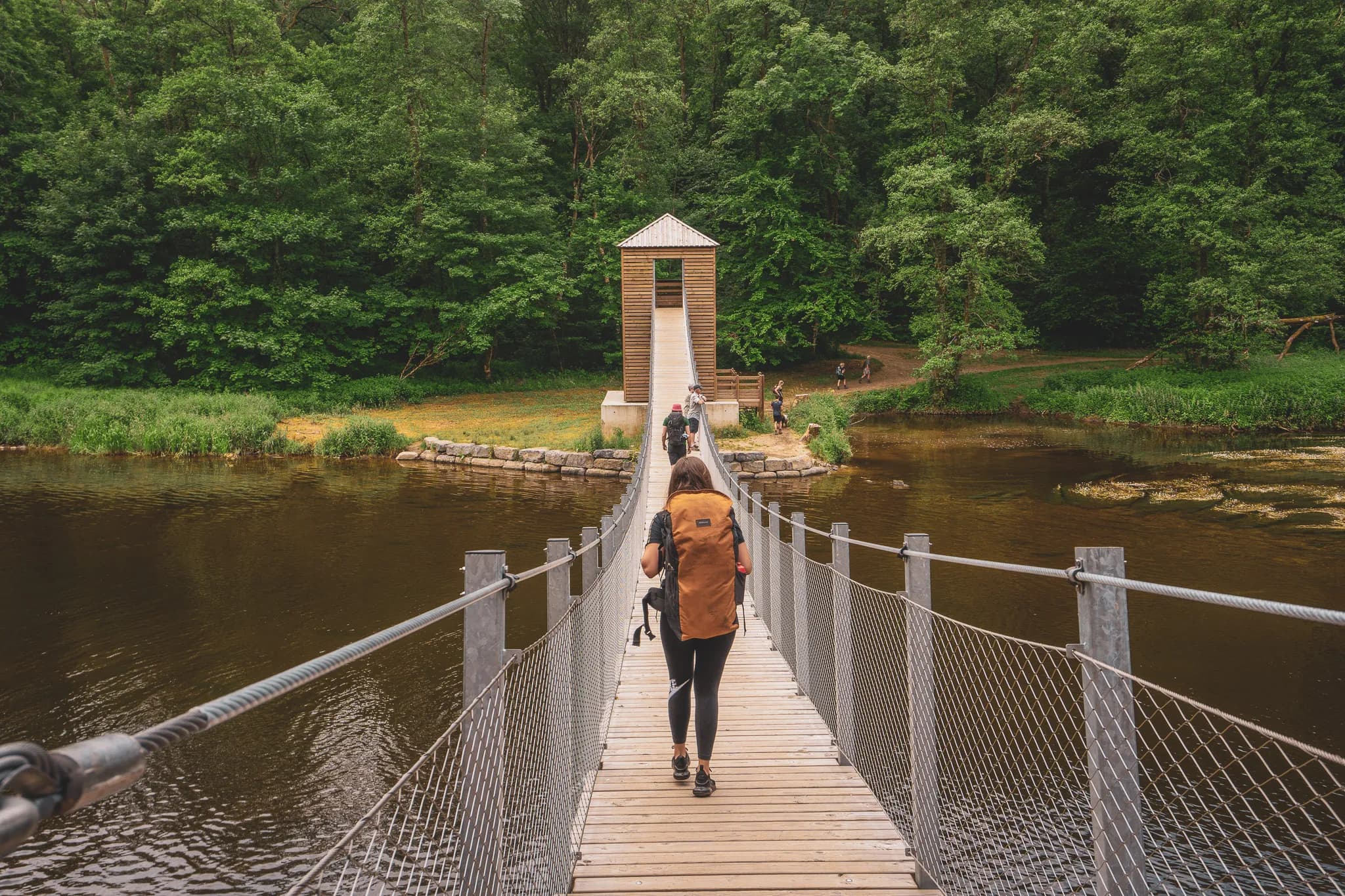 Een houten hangbrug overspant een kalme rivier, omgeven door dicht groen. In het midden van de brug beweegt een persoon met een grote rugzak vooruit, terwijl verschillende andere mensen vlakbij op de oever staan, sommigen met een