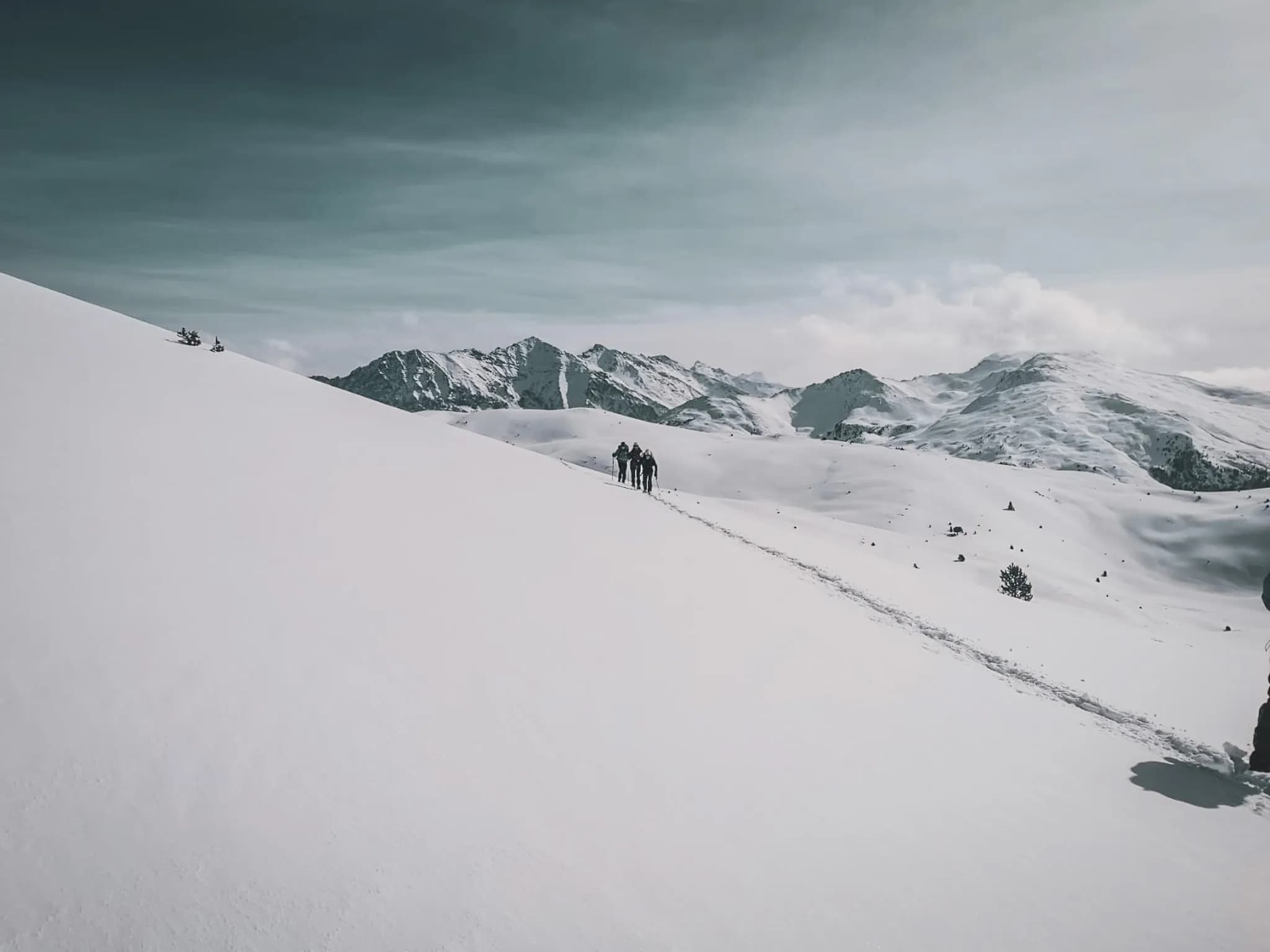 A snow-covered alpine landscape with imposing mountains in the background. The scene is dominated by a vast field of virgin snow, dotted with small rocks. On the other side, a group of hikers