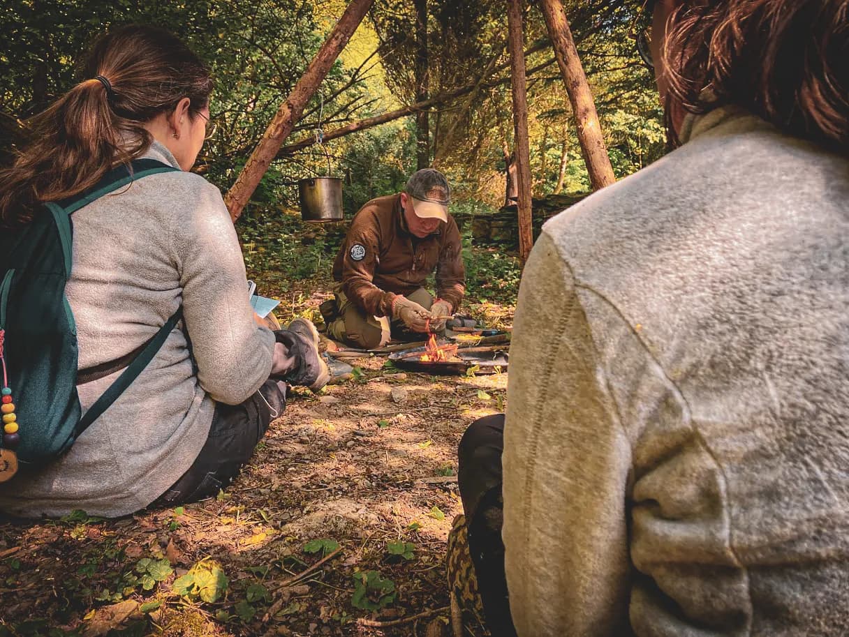 Een groep in het hart van de natuur, leren overleven rond een vuur, in de serene sfeer van de Ardennen.