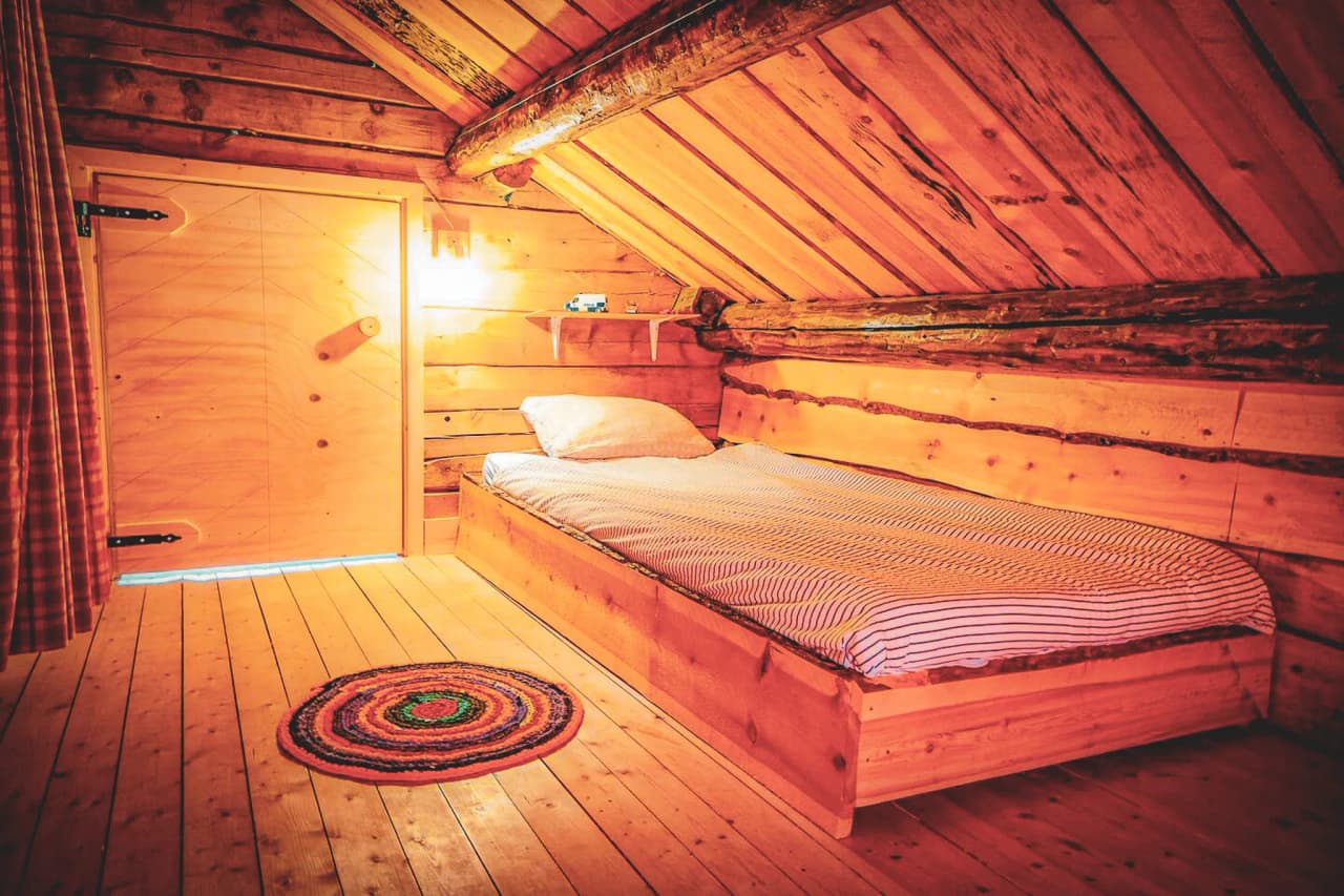 A cosy wooden bedroom with natural wood plank walls and a sloping ceiling. A wooden bed is set against the wall, covered with a white and grey striped duvet. On the floor, a