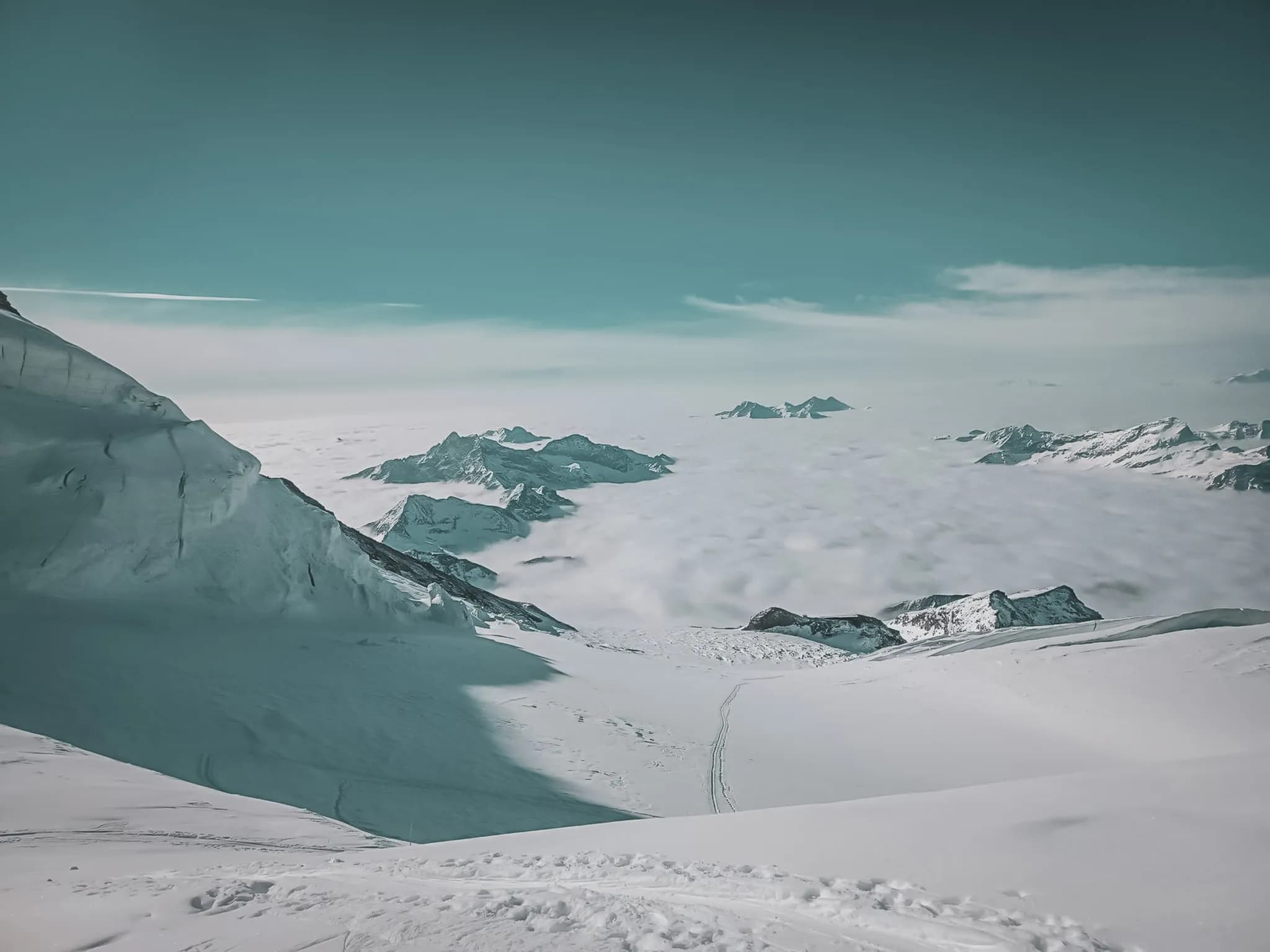 Een spectaculair Alpenlandschap, met majestueuze bergen omringd door sneeuw. Daarboven een zachte, licht bewolkte lucht, terwijl zich daaronder een zee van wolken uitstrekt zover het oog reikt,