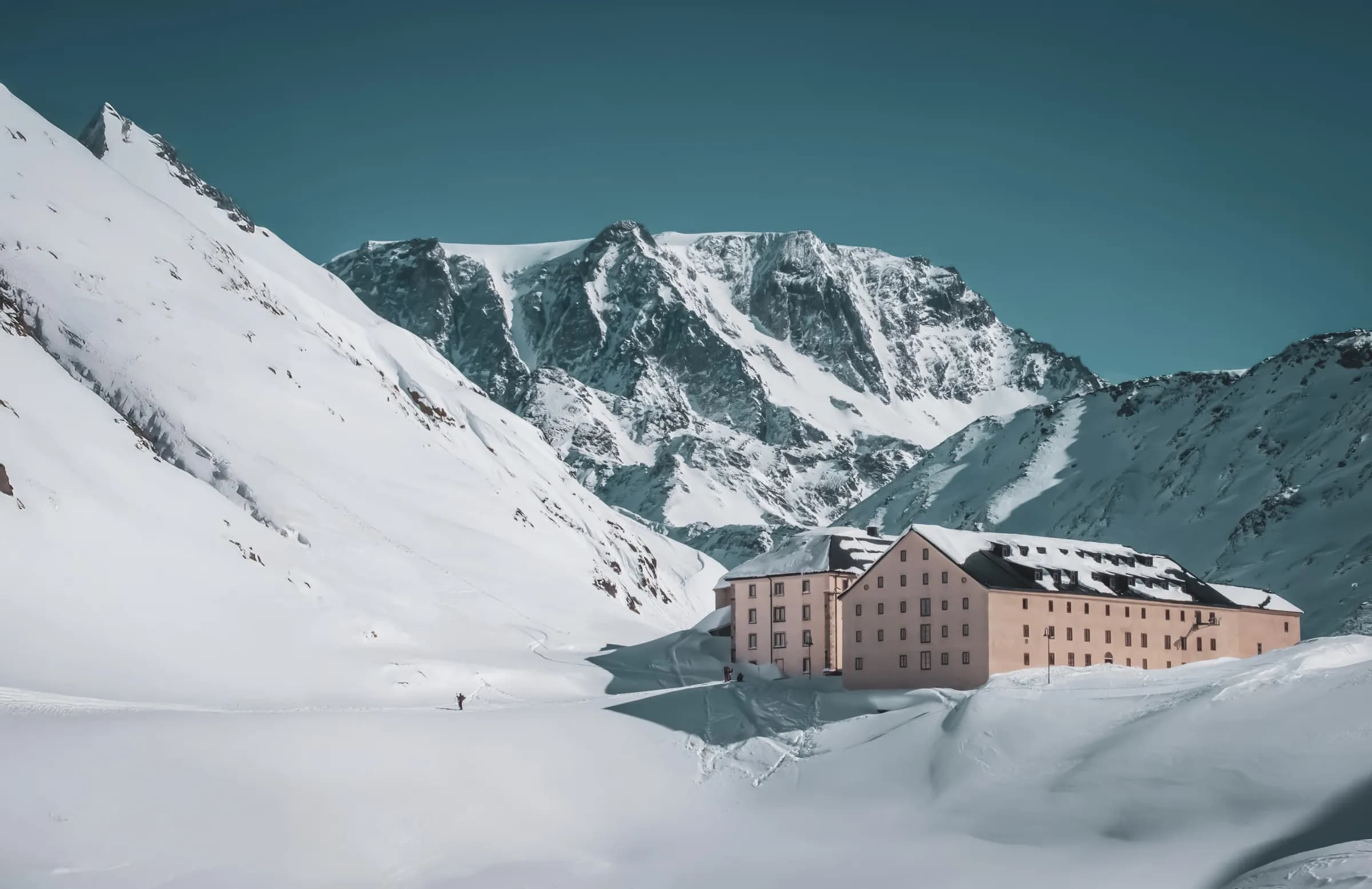 A chalet-style building is framed by a snow-covered mountain landscape. The imposing peaks, adorned with glaciers, rise majestically in the background against a clear blue sky. The architecture