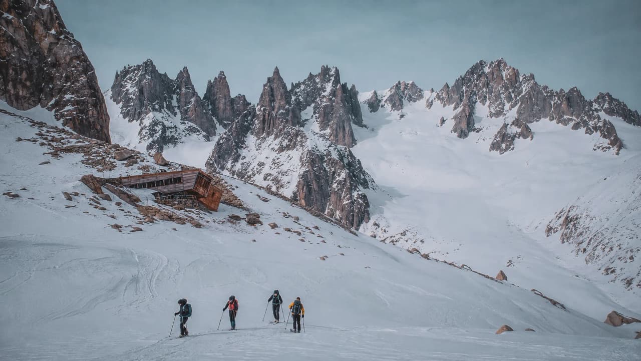 Paysage alpin enneigé, randonneurs en ski devant des pics majestueux et un chalet abandonné.