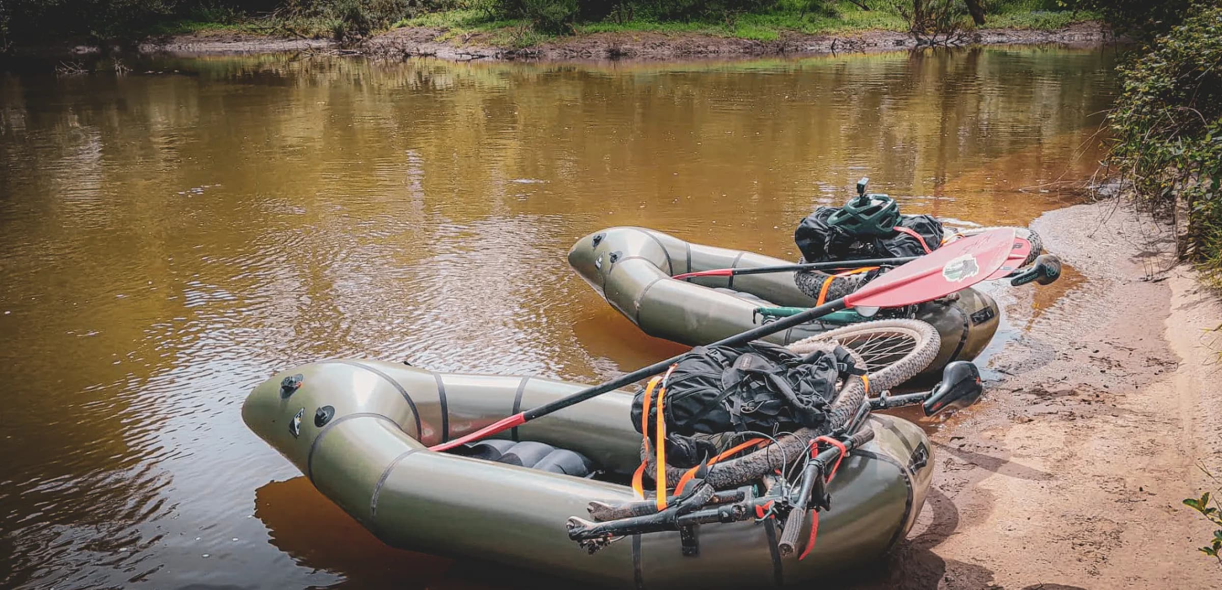 Two inflatable canoes are anchored on a quiet bank, with matching hiking and mountain biking equipment alongside. The canoes are lined with black sacks, dusty bicycles, and a few other things.