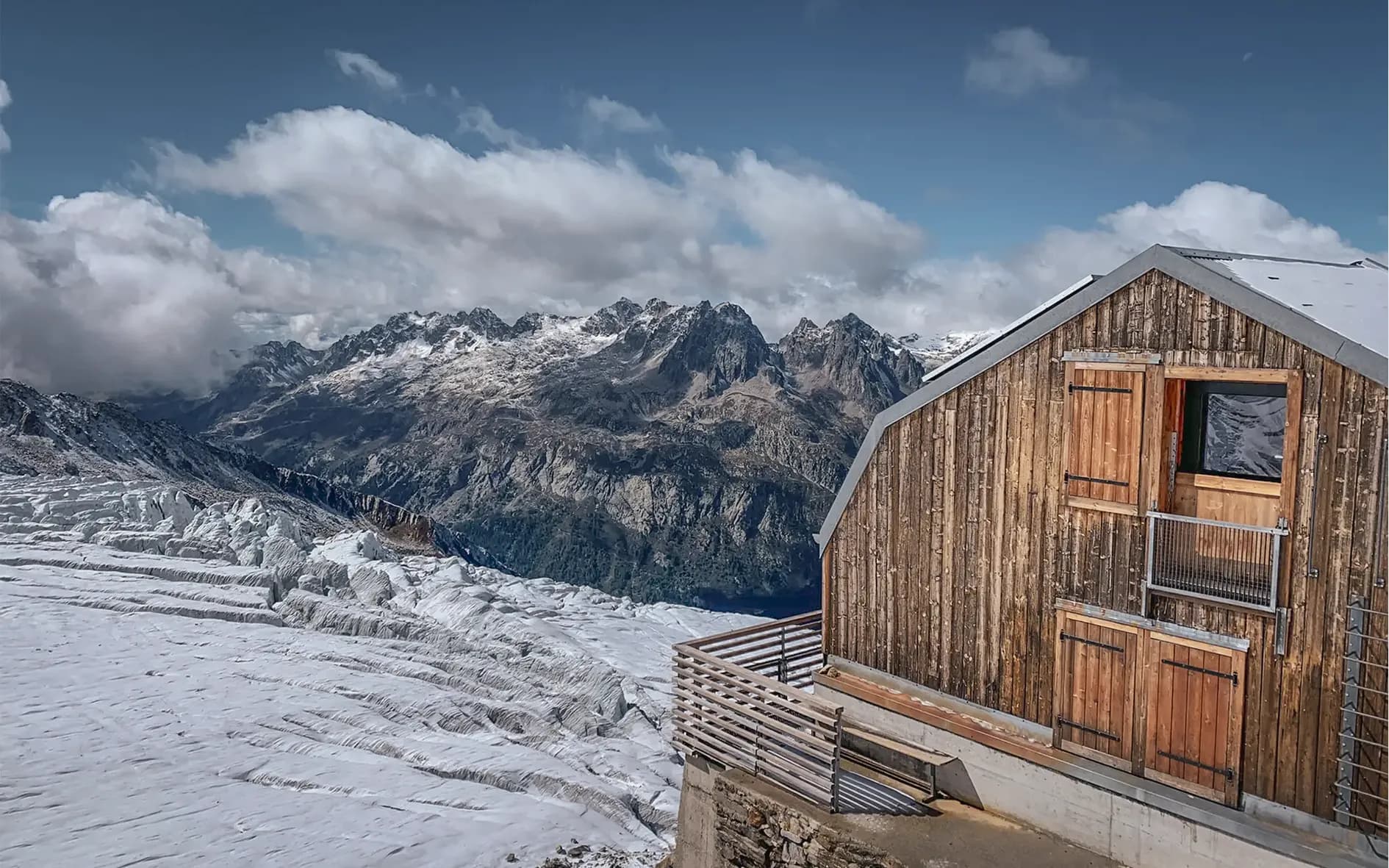 A wooden chalet set in steep terrain with panoramic views of majestic mountains. The peaks are covered in snow and rise up under a partly cloudy sky. The glacier below
